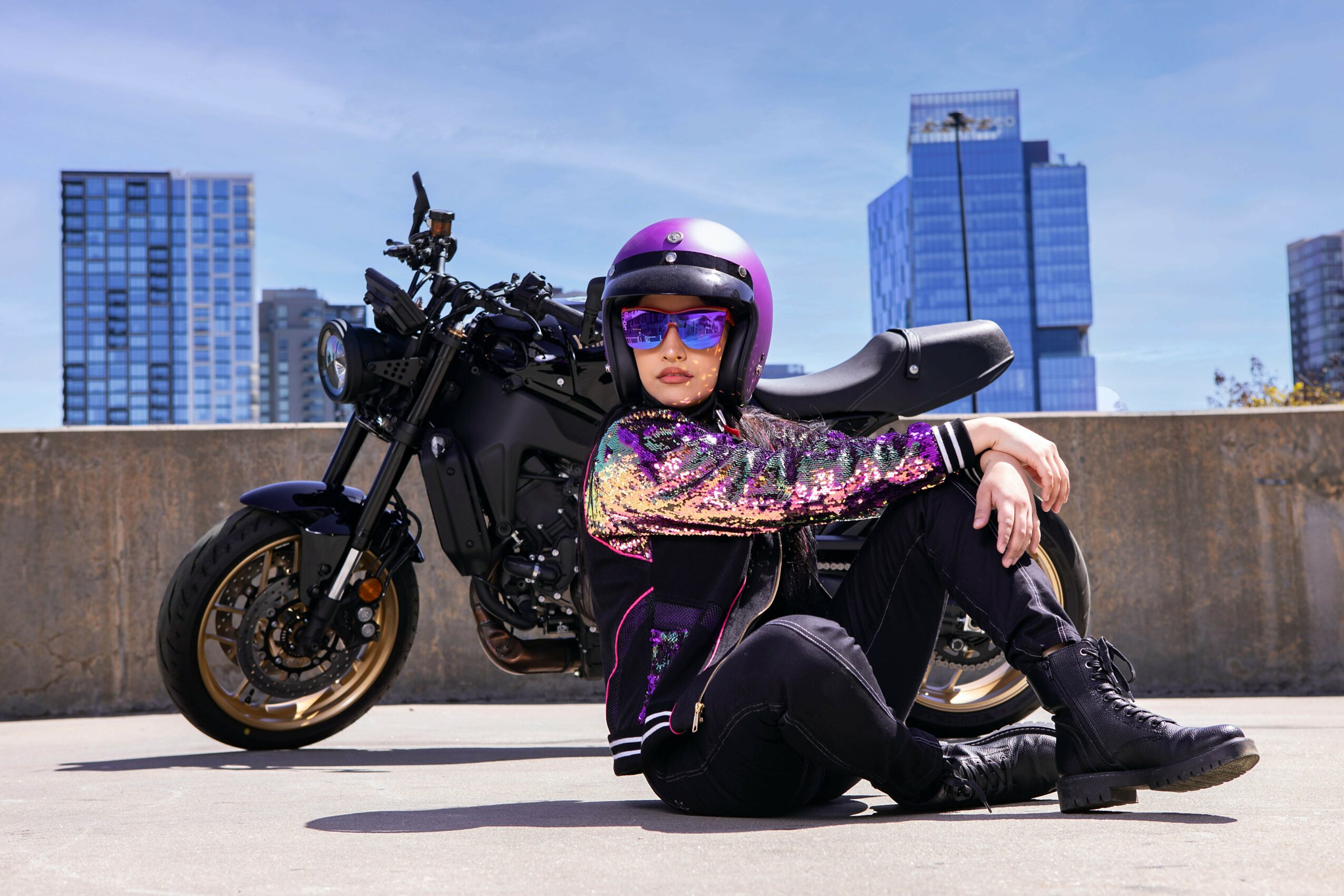 Person sitting on ground next to a black motorcycle with city buildings in background, wearing a helmet and sunglasses.
