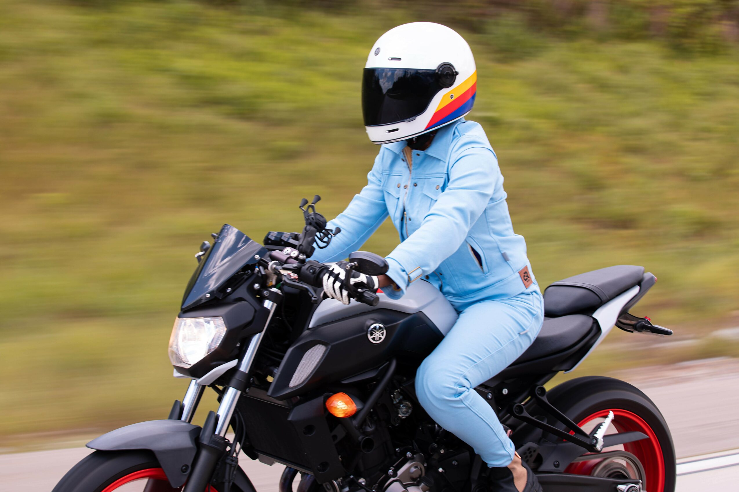 Person riding a black motorcycle with a white helmet, wearing a light blue outfit, on a blurred outdoor background.
