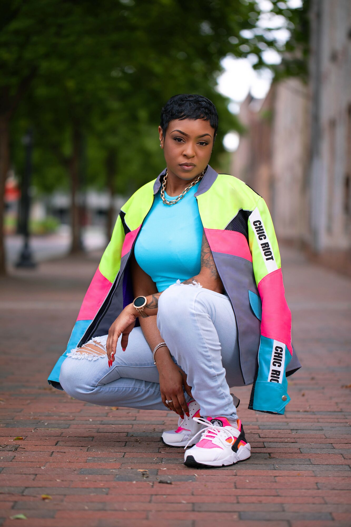Woman crouching on brick sidewalk, wearing colorful jacket, blue top, white pants, and sneakers, with trees and buildings in background.