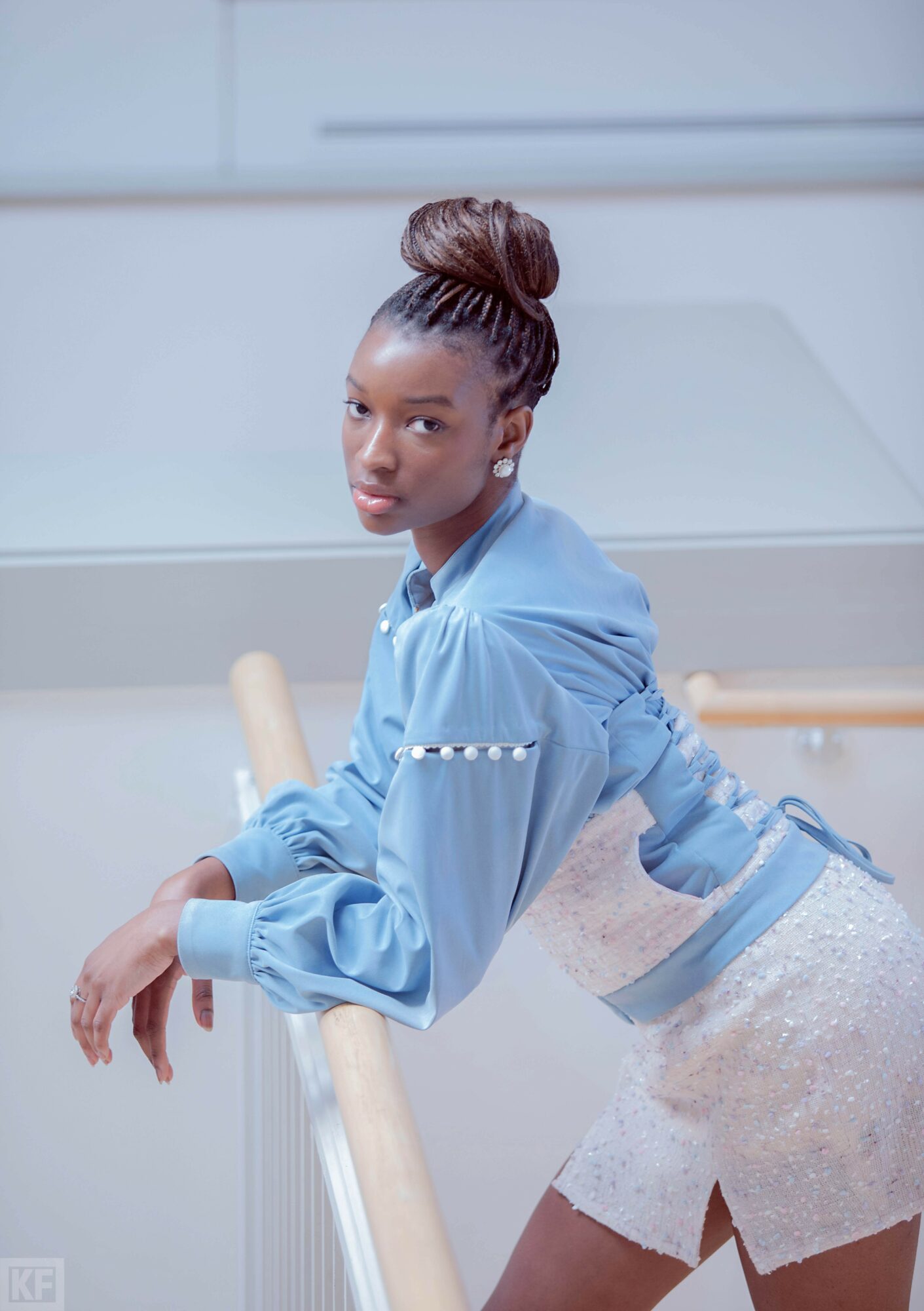Young woman with braided hair in a blue blouse and white skirt leaning on a ballet barre in a dance studio.