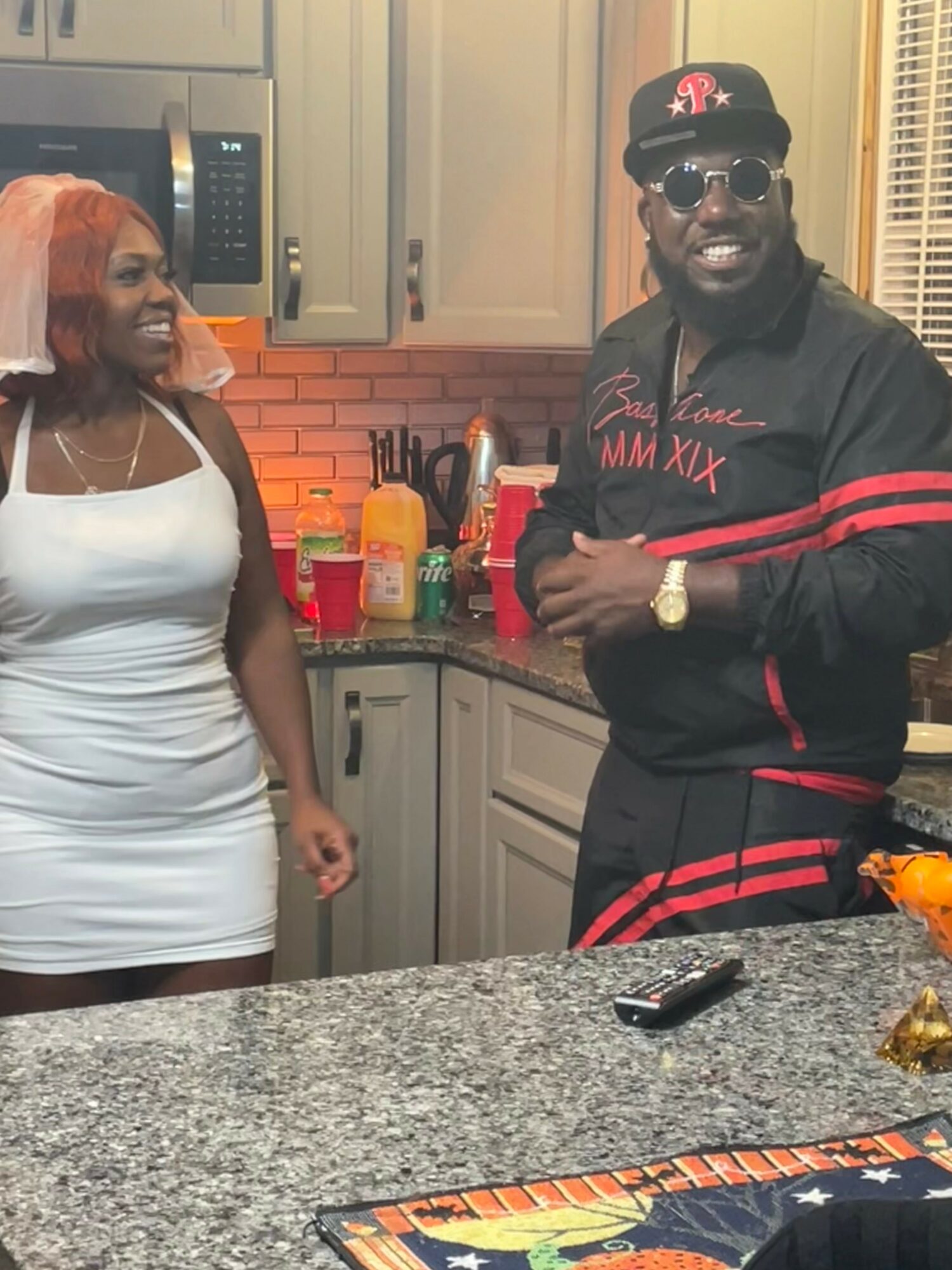 Two people in a kitchen, smiling and talking, with various items on the counter and cabinets in the background.