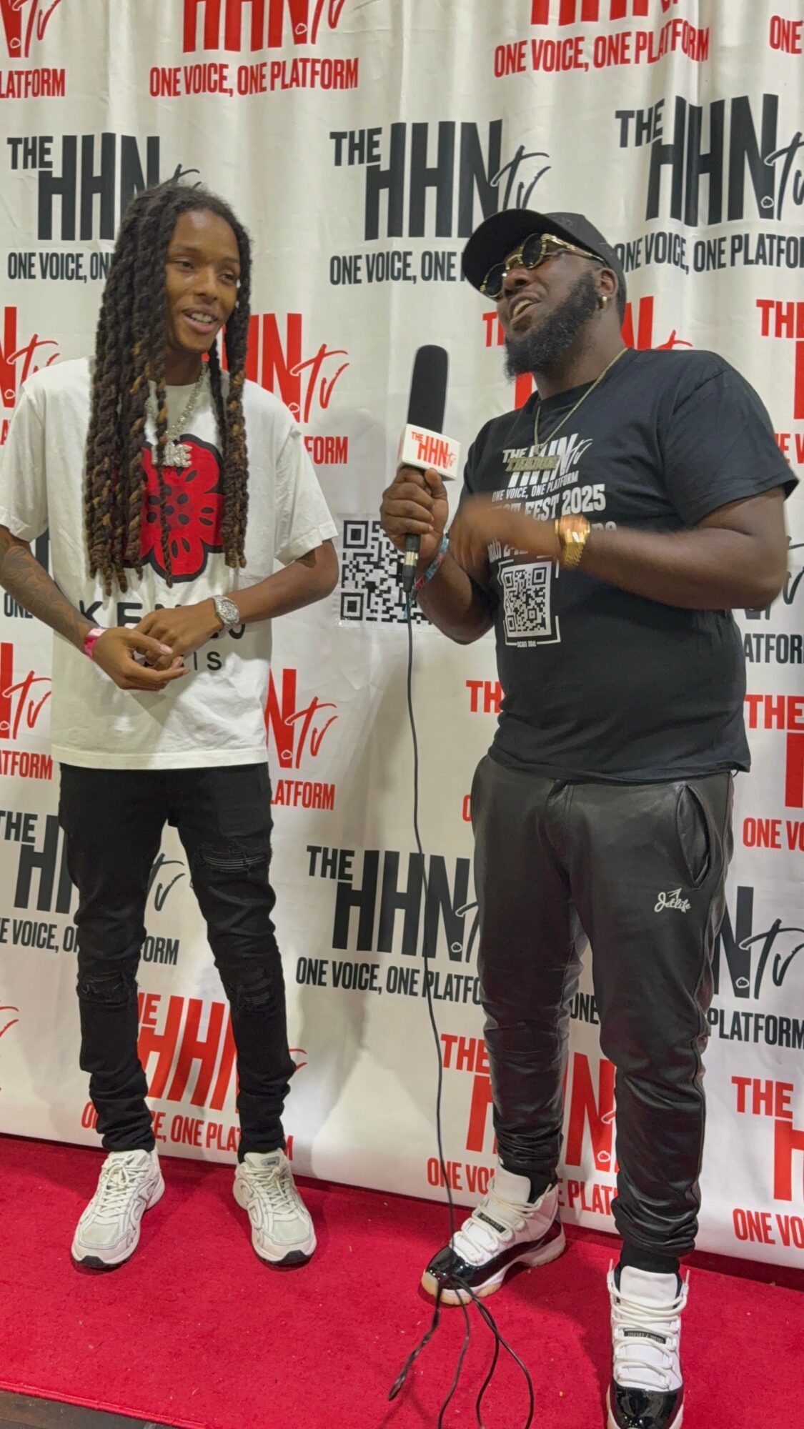 Two people standing on a red carpet in front of a backdrop with logos, one holding a microphone, both smiling.