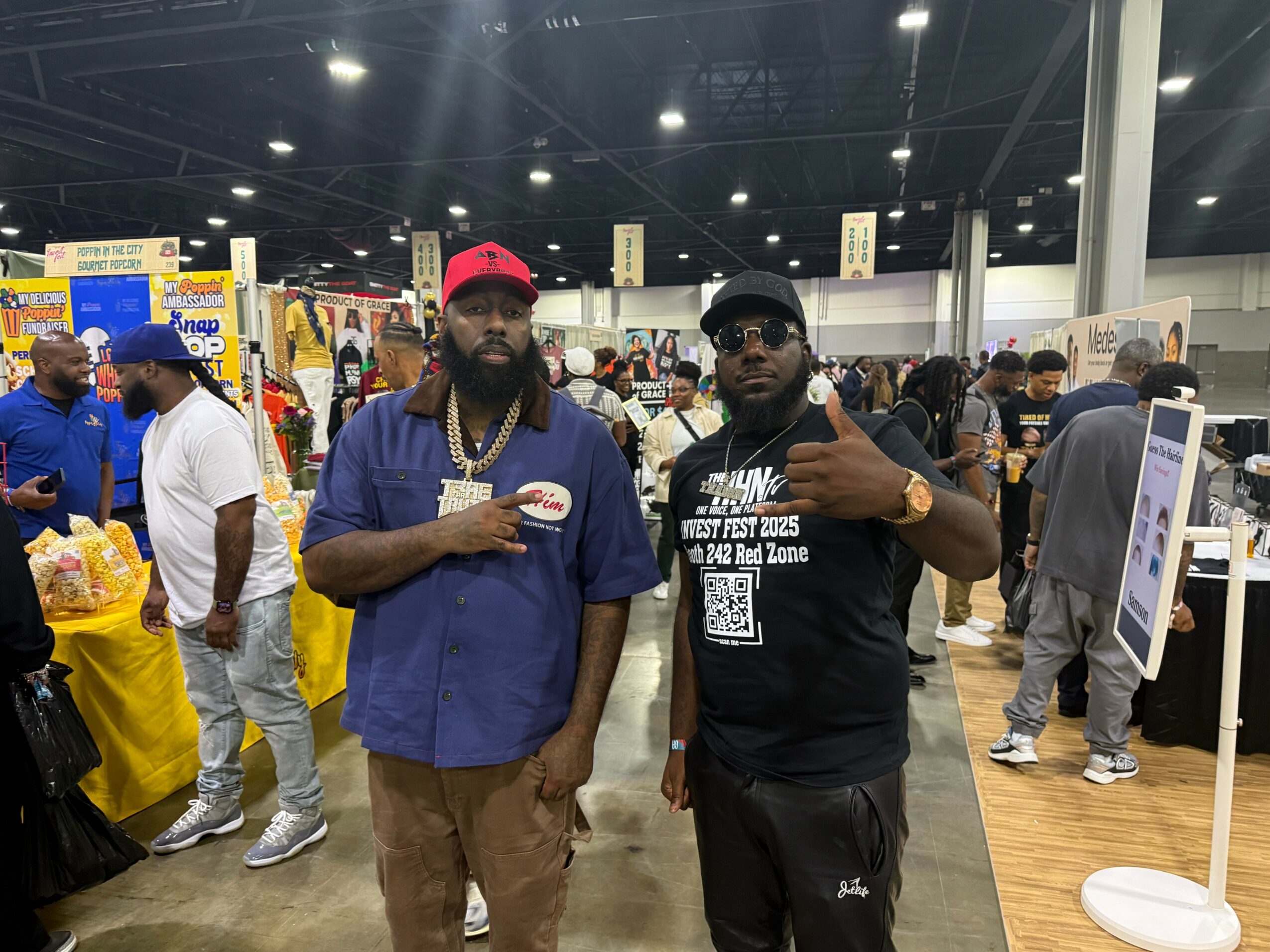 Two men standing in a busy indoor event, one wearing a red cap and blue shirt, the other in black, surrounded by people and booths.