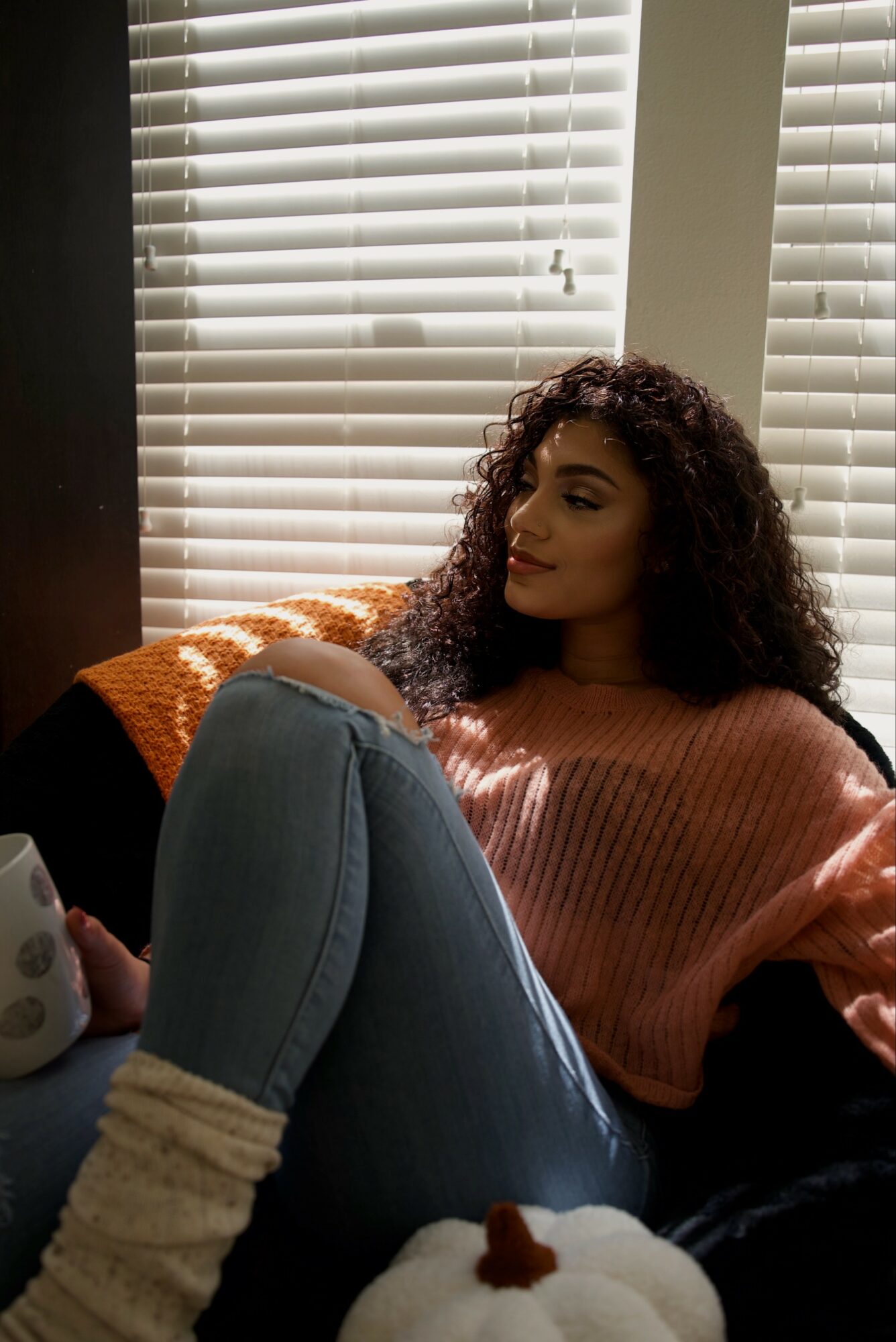 Young woman with curly hair sitting by window with blinds, wearing a pink sweater and jeans, holding a mug.
