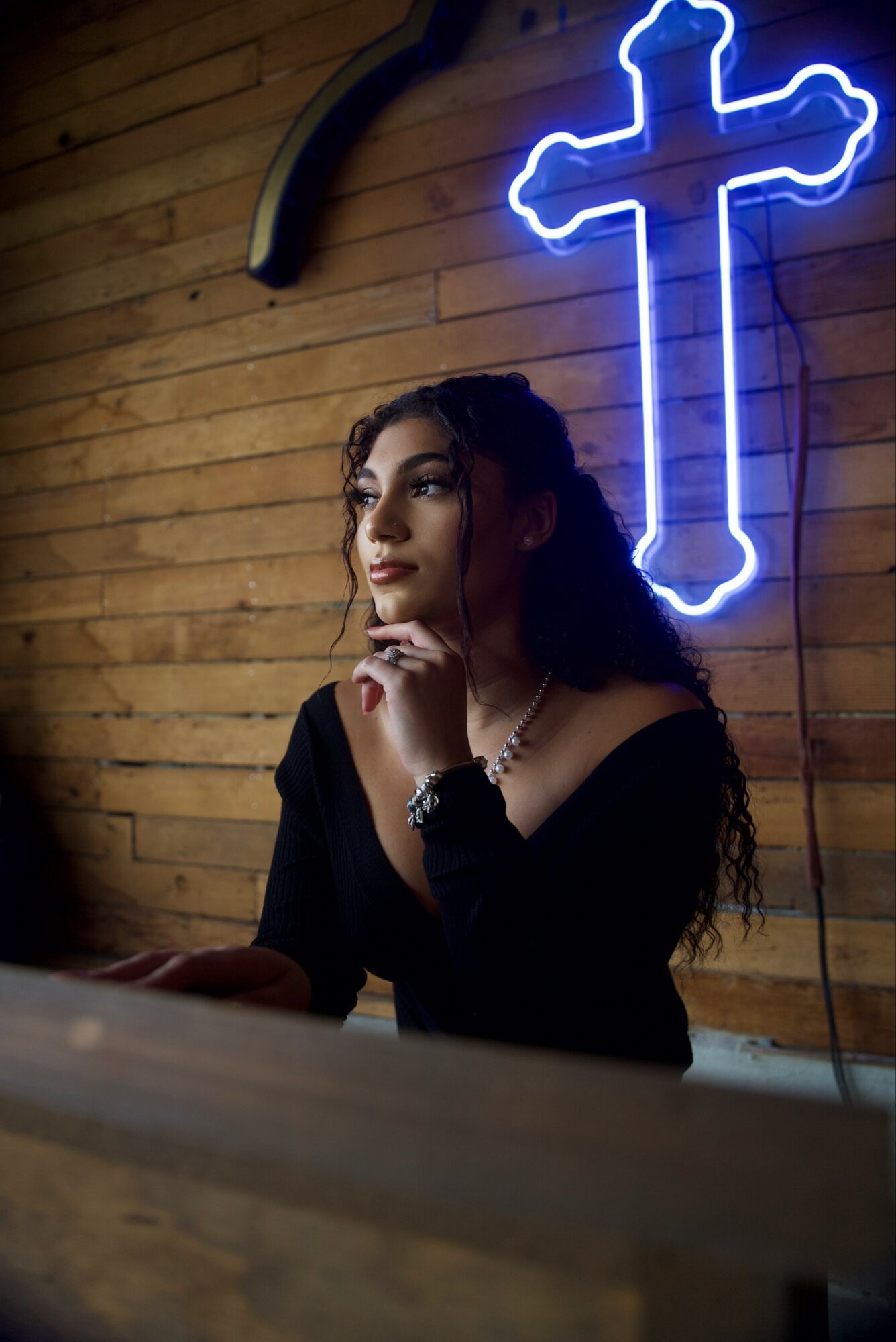 Woman with dark hair and makeup, wearing black, sitting near a wooden wall with a neon cross light behind her.