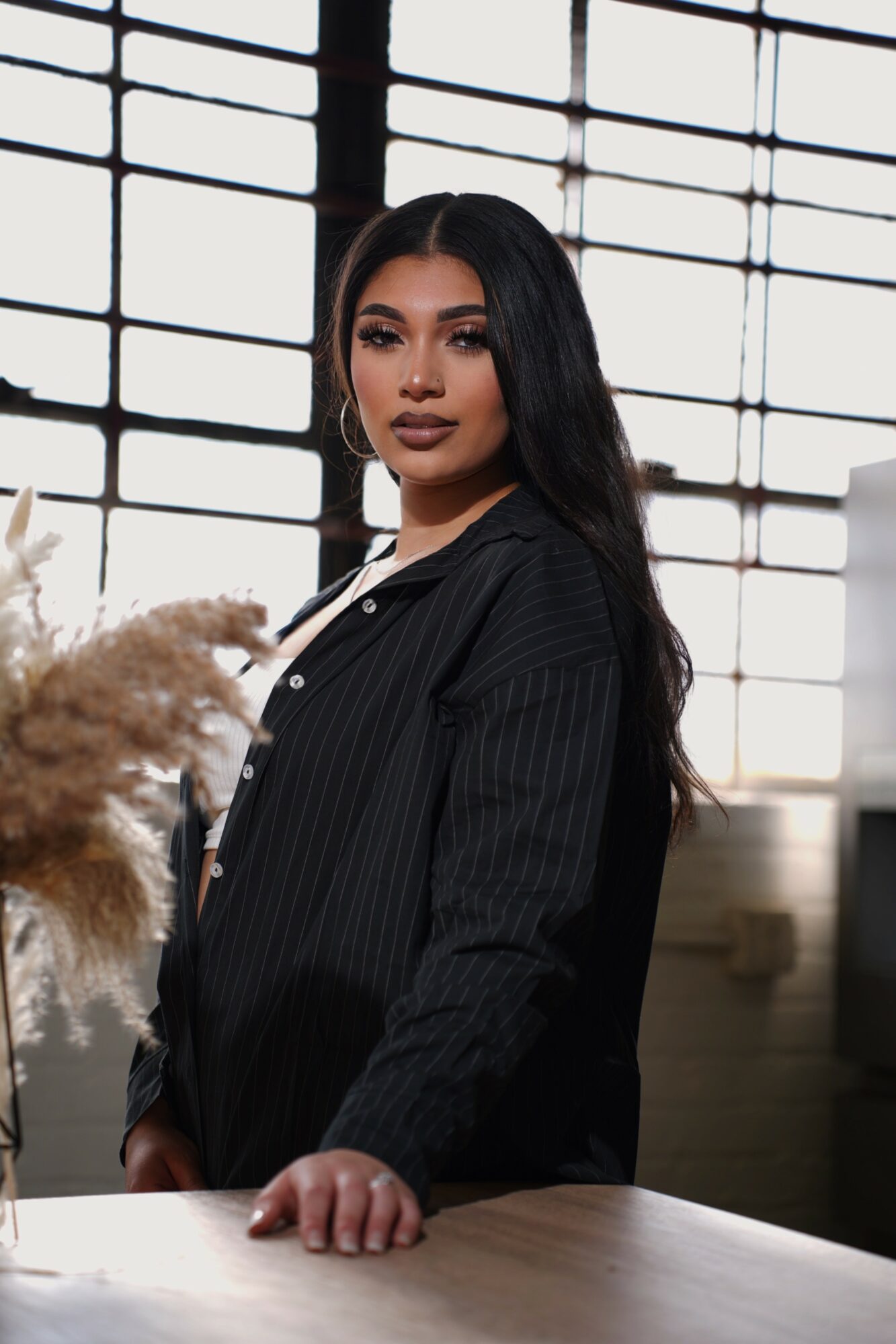 Woman with long dark hair and makeup, wearing a black pinstripe shirt, standing indoors near a table with a window background.