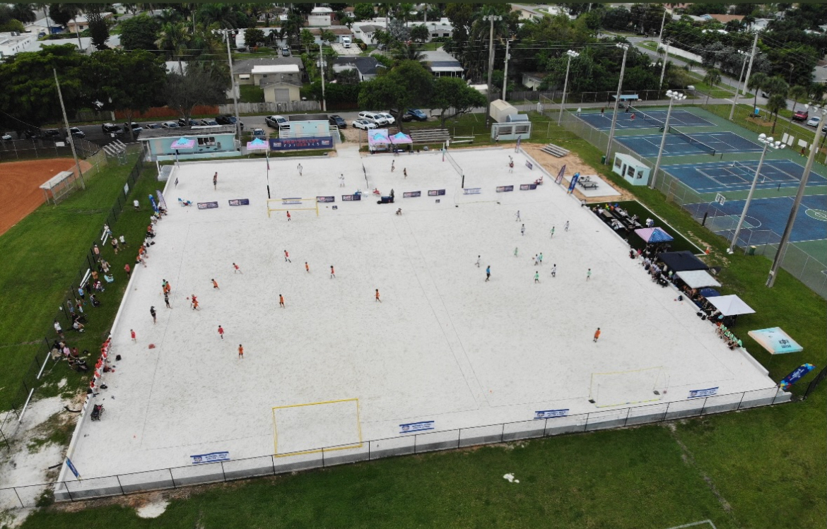 Aerial view of a sports field with tennis courts and surrounding area, with multiple people on the field and courts.