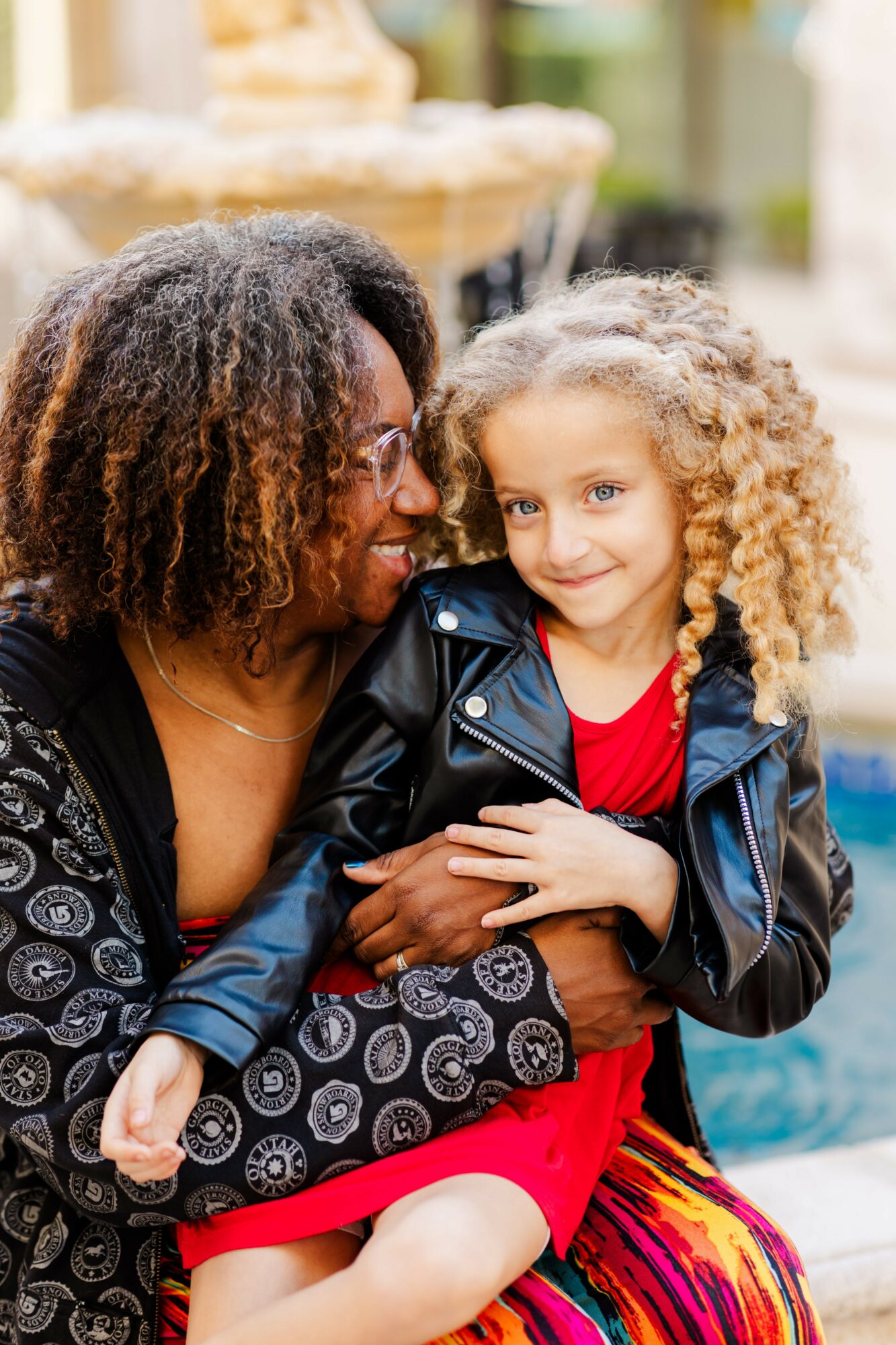 Adult woman and young girl with curly hair smiling and embracing outdoors, blurred background, colorful clothing.