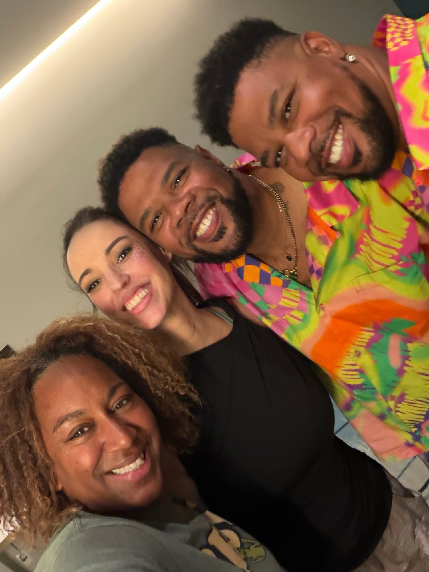 Four people smiling closely together indoors, with a plain wall background.