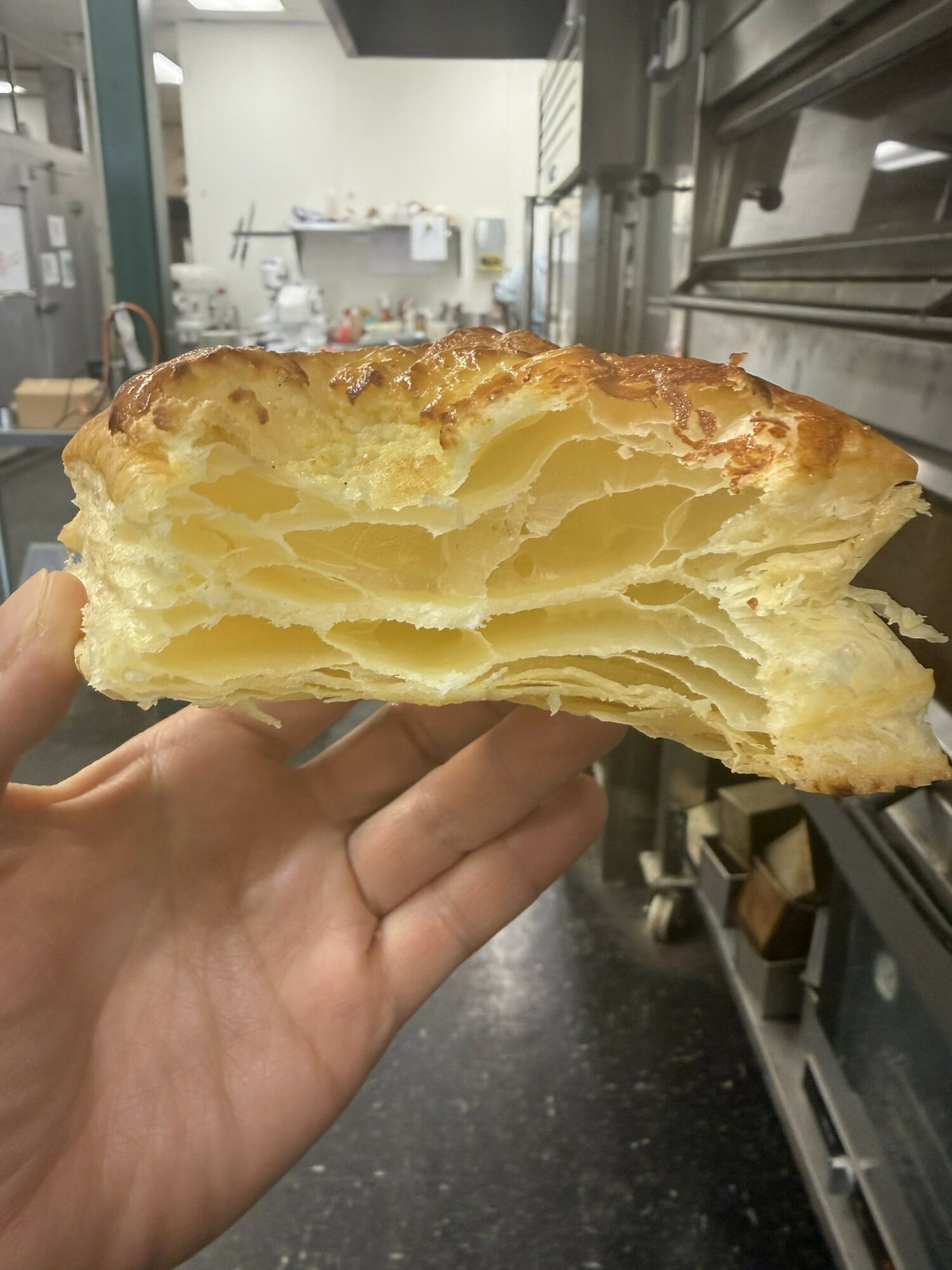 A hand holding a flaky, layered pastry with a golden-brown crust, inside a commercial kitchen.