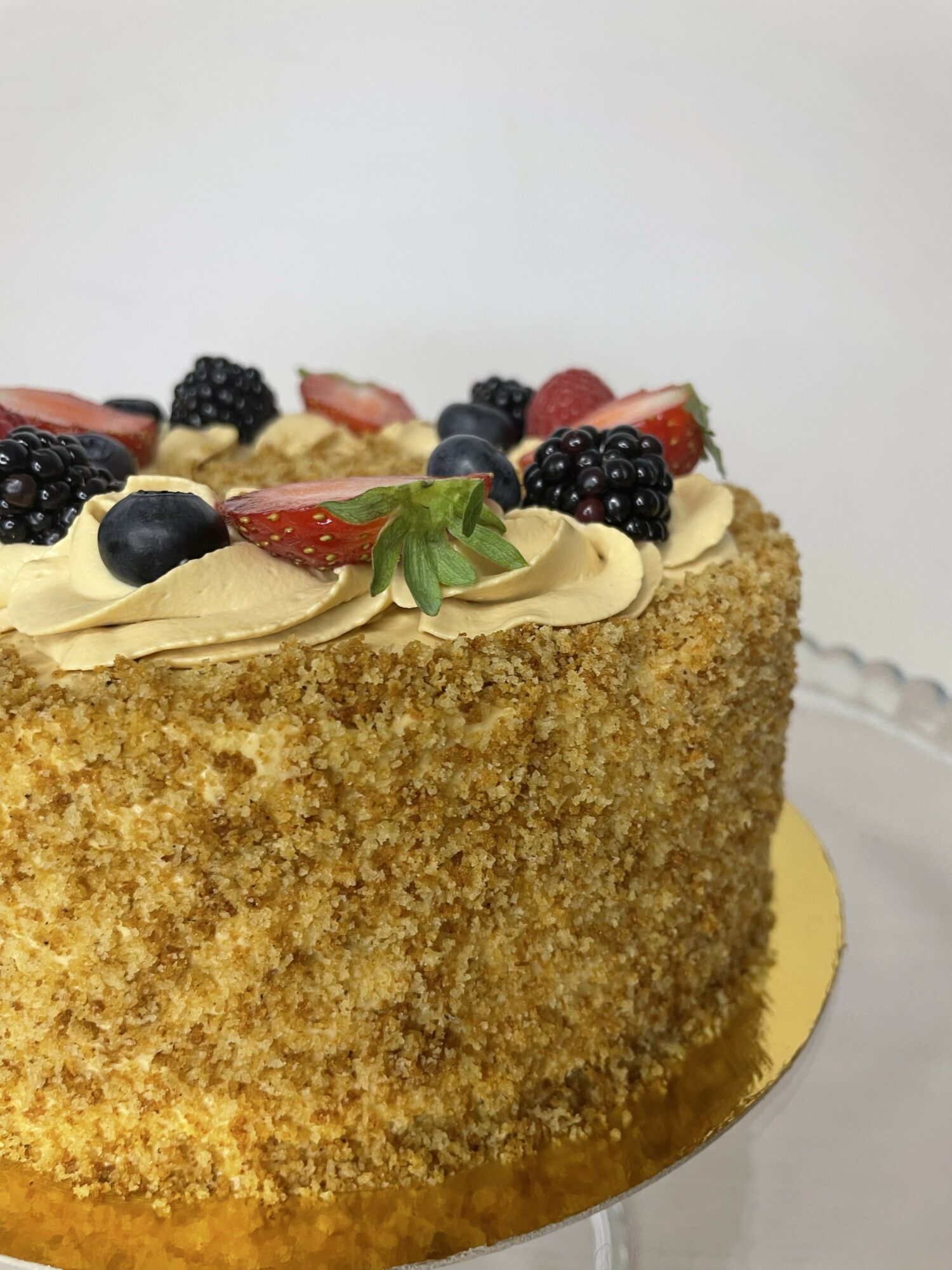 Cake with a crumb coating, topped with mixed berries and cream, on a gold base against a plain background.