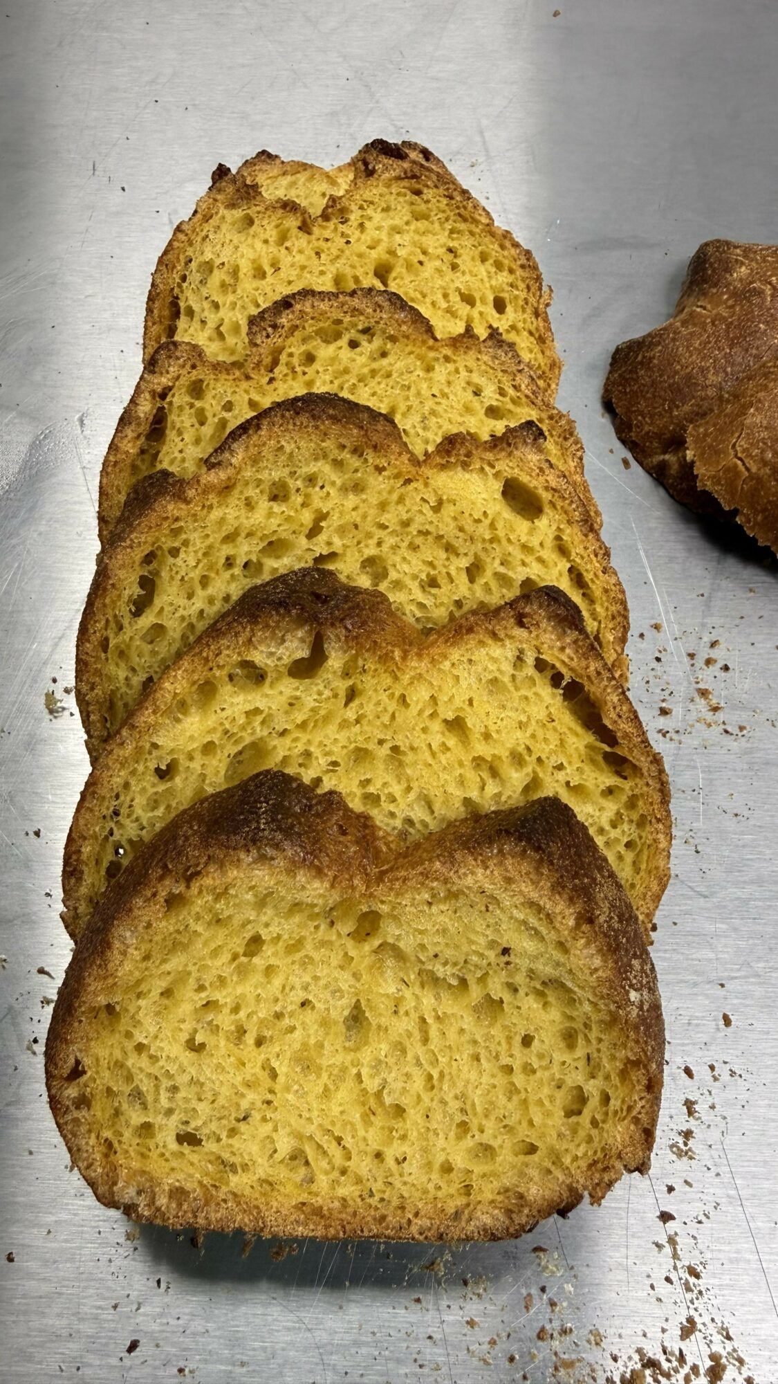 Five slices of yellow cake with a brown crust stacked on a surface, with a partially visible baked item nearby.