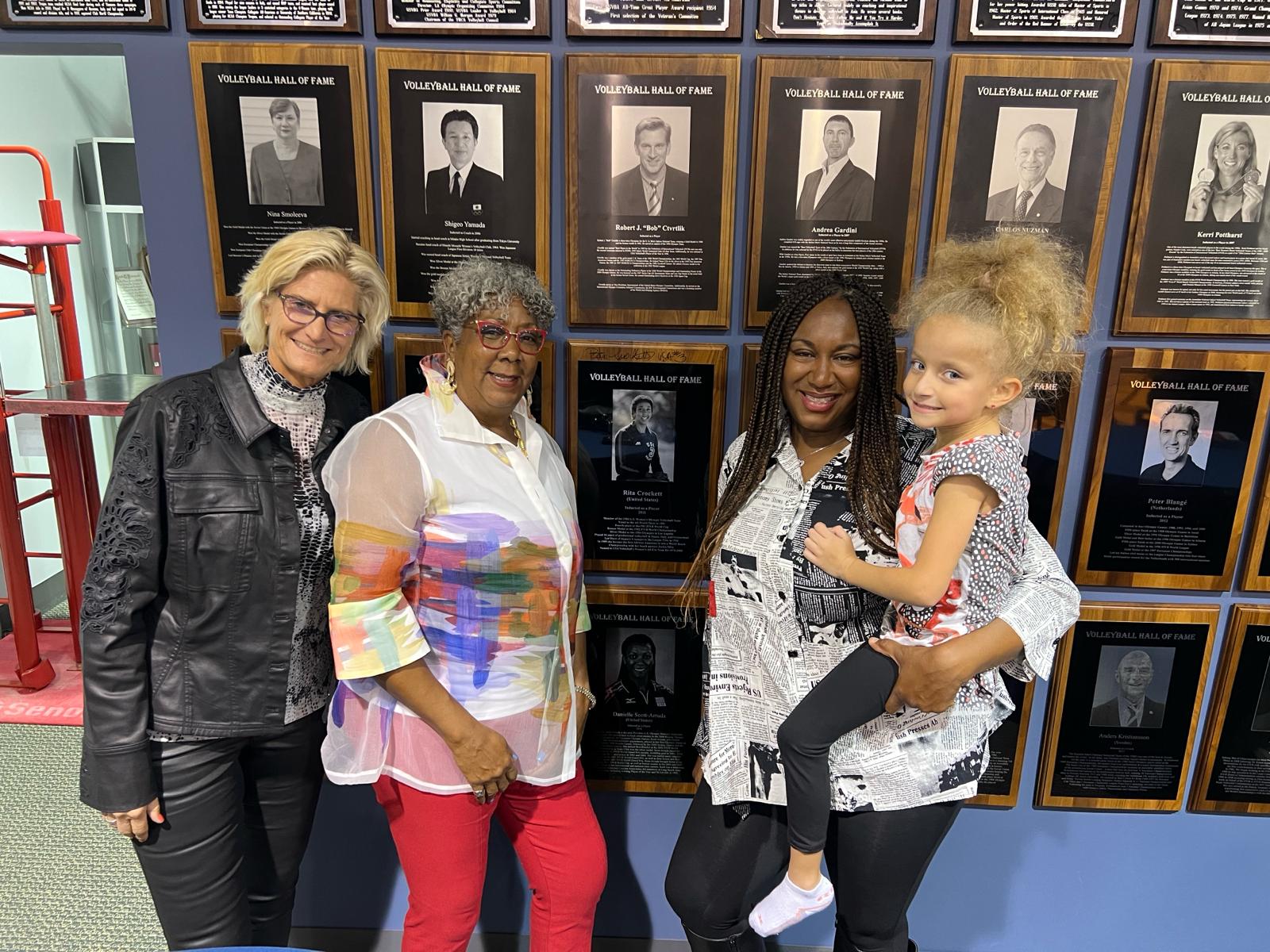 Four women standing in front of a wall with framed photographs and descriptions, smiling at the camera.