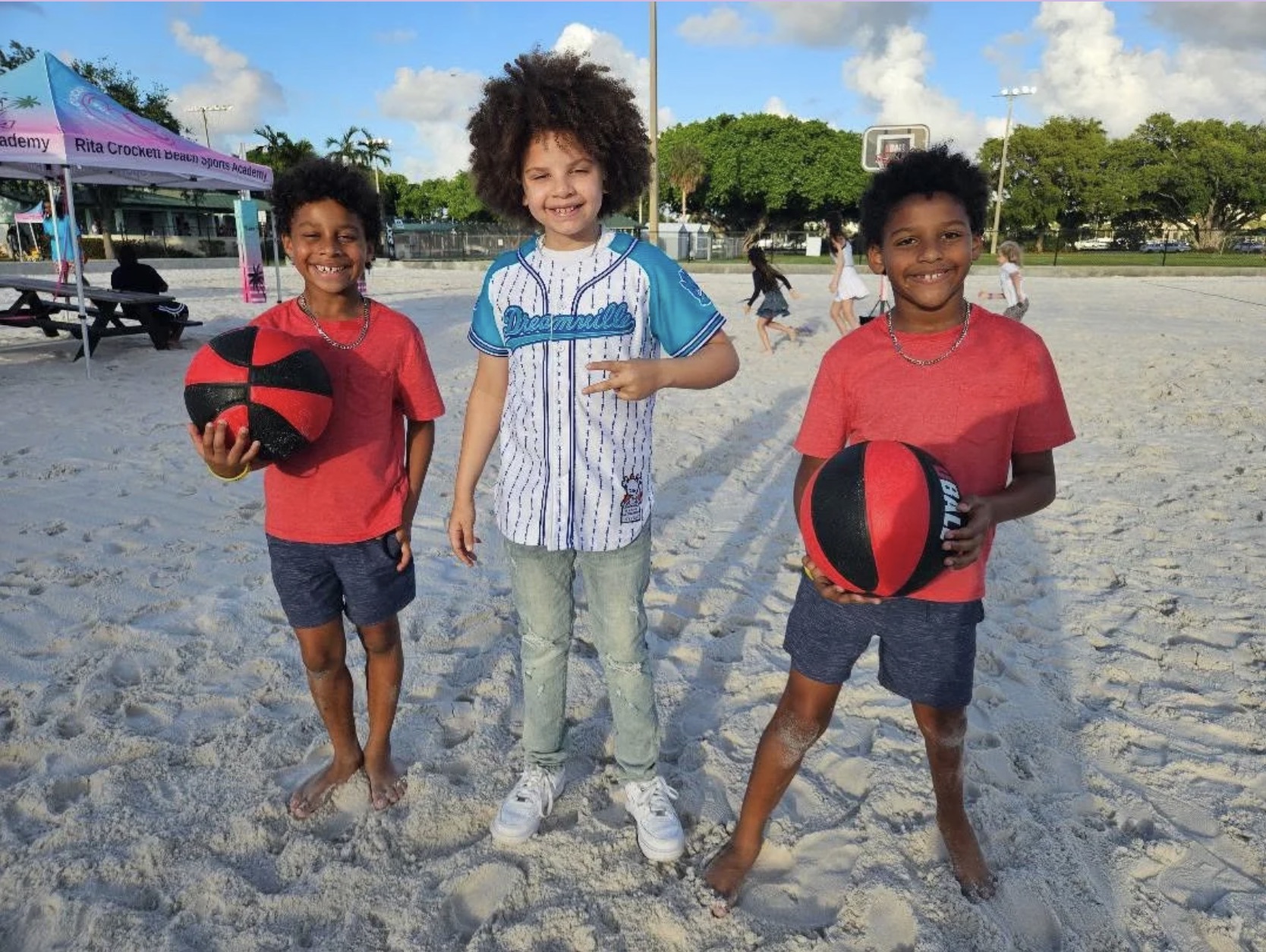 Three children standing on a sandy outdoor court holding basketballs, with trees and a blue sky in the background.