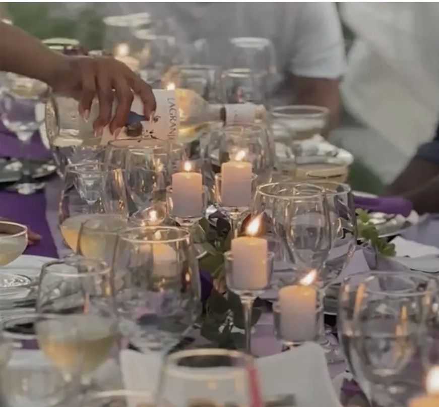 Person pouring wine at a table with candles, glasses, and tableware, outdoors during daytime.