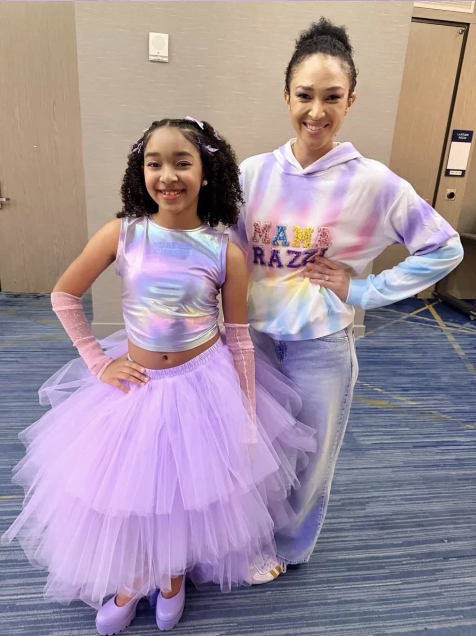 Two smiling females, one young girl in a pastel rainbow top and purple tutu, and an adult woman in a tie-dye hoodie and jeans, standing indoors.