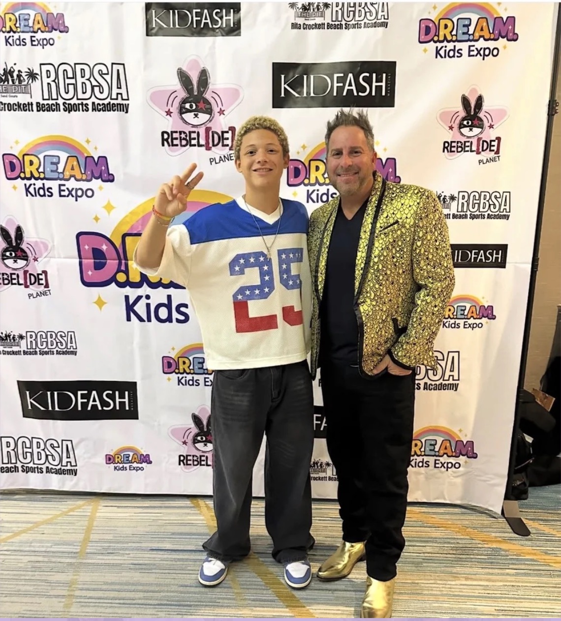 Two people standing in front of a backdrop with event logos, smiling and making peace signs.