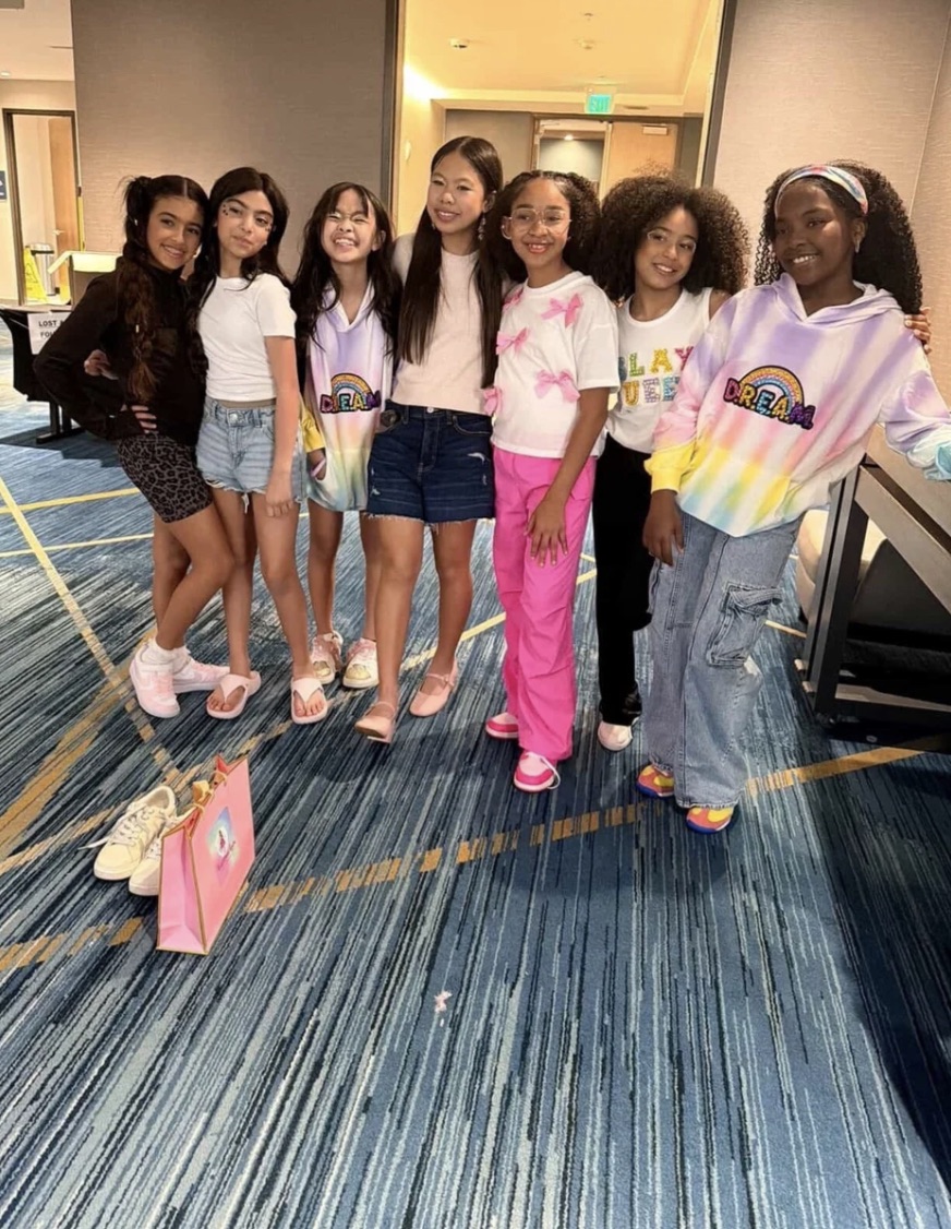 Group of seven young girls standing together indoors, smiling, with a blue carpeted floor and a pink bag on the floor.