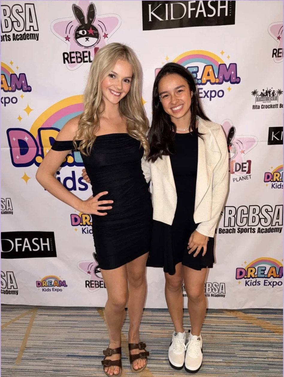 Two smiling girls standing in front of a colorful event backdrop, one with blonde hair in a black dress, the other with dark hair in a white jacket and black dress, both wearing sneakers and sandals.