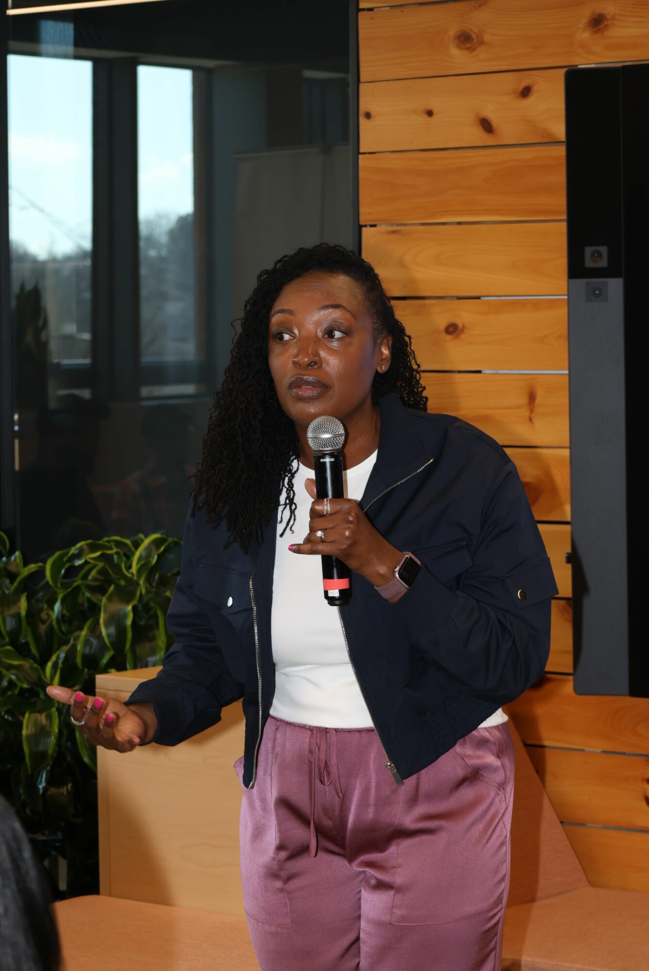 Woman speaking into a microphone, gesturing with her left hand, standing indoors with wooden wall and window behind her.