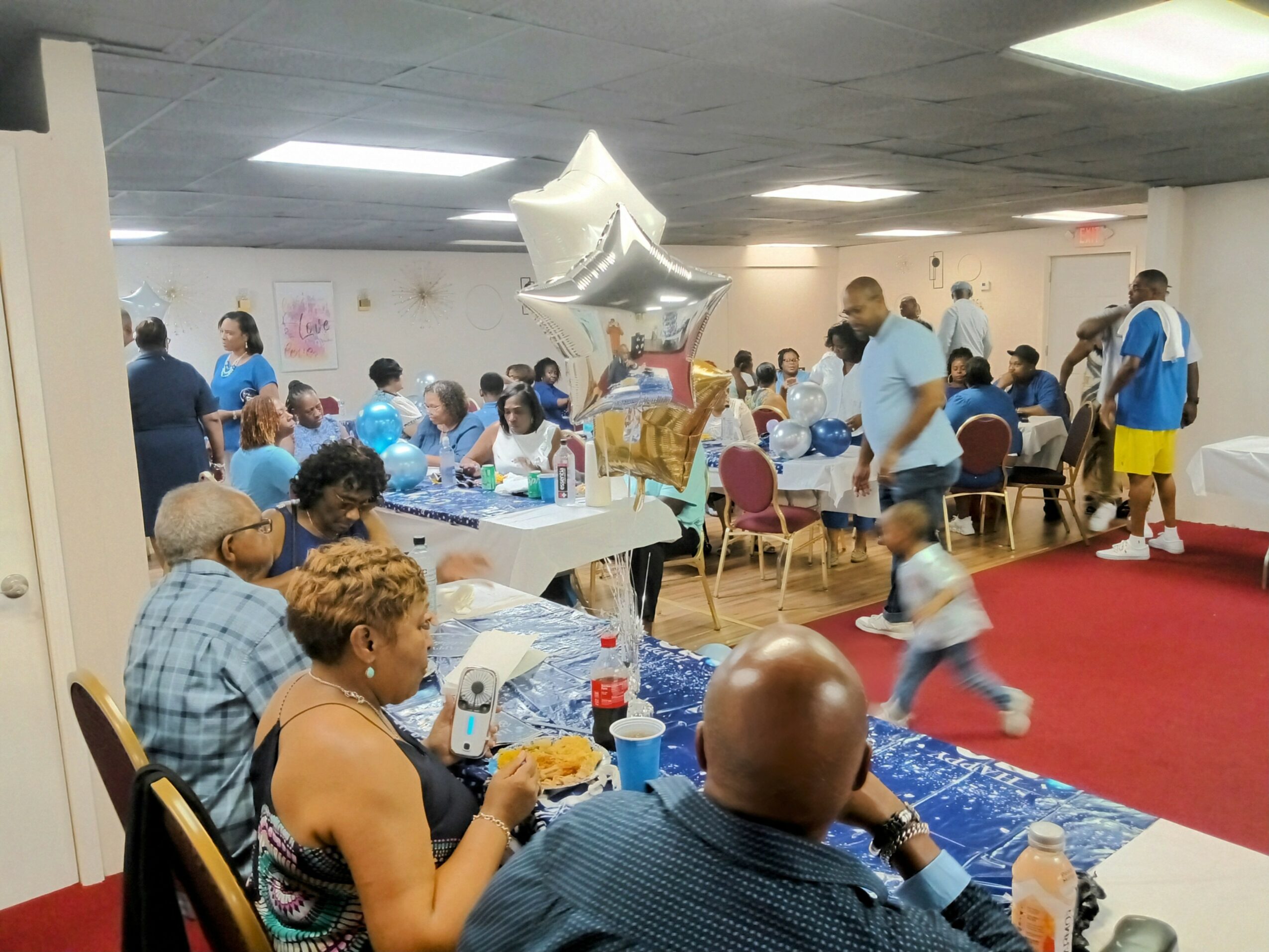 People gathered in a room with tables, balloons, and children, celebrating an event with food and decorations.