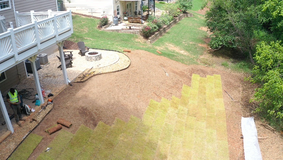 Yard with partially laid yellow and green grass, trees, and a house with a deck, construction in progress.