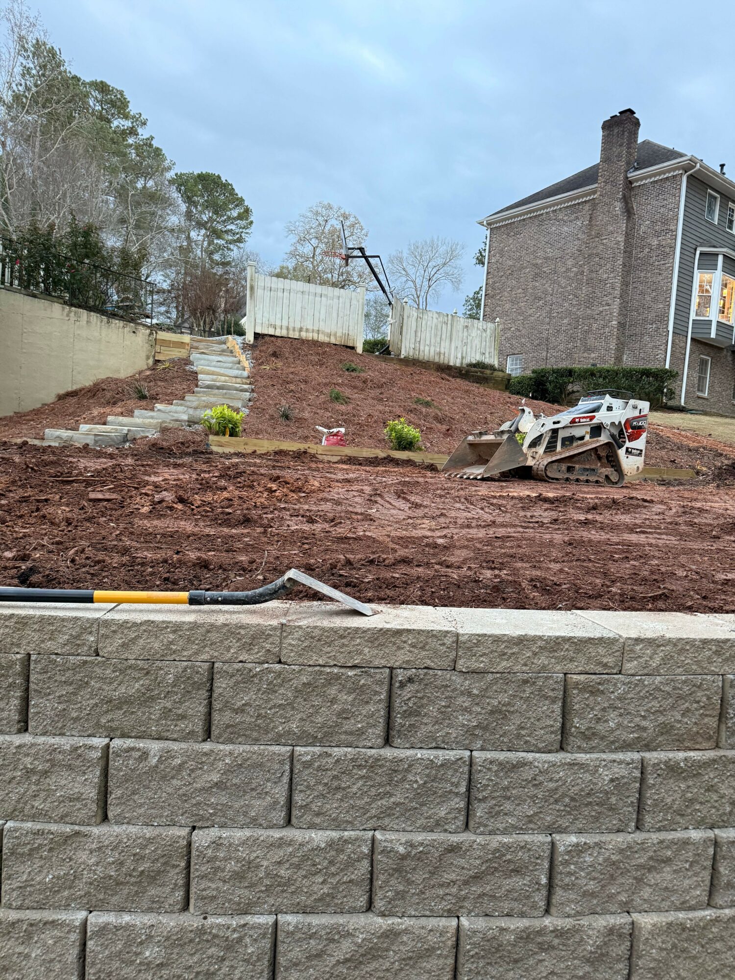 Construction site with dirt, paving stones, and a wheelbarrow, next to a brick wall and residential buildings in background.