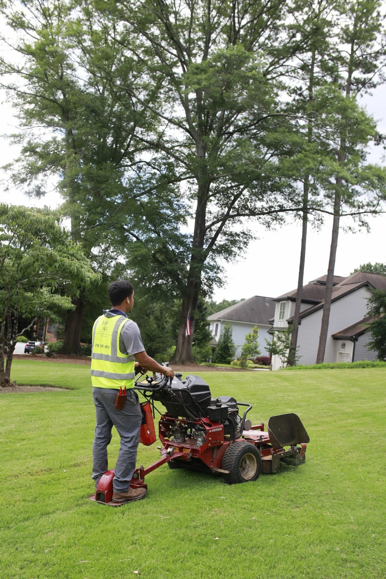Person operating a lawn mower on a grassy area with trees and houses in the background.
