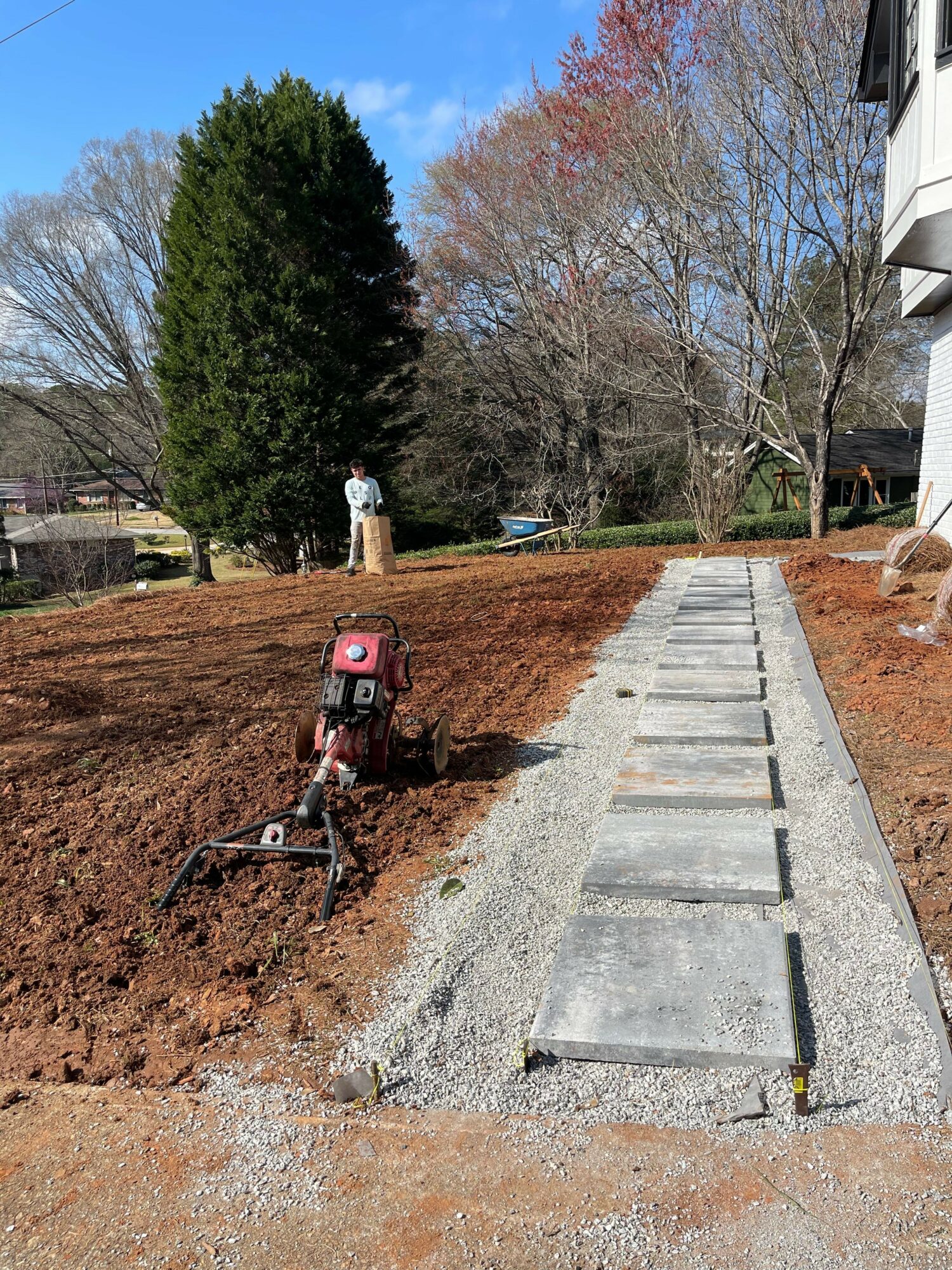 Concrete pathway with rectangular stepping stones in a garden, trees, and a person in the background.