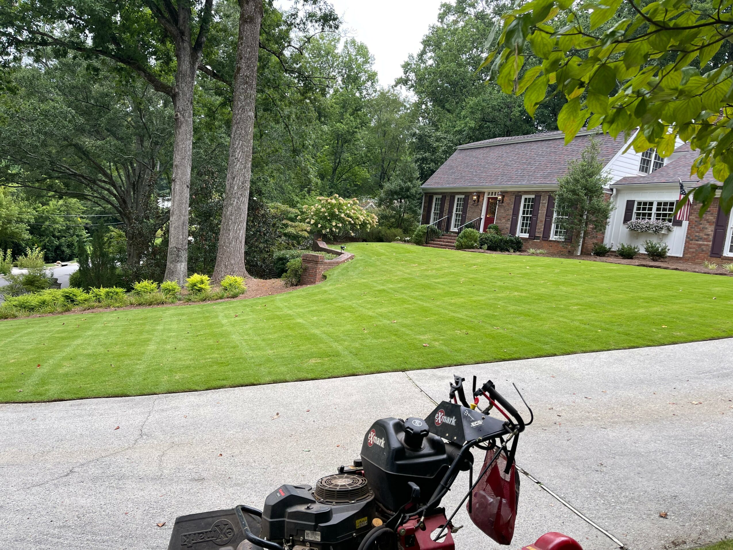 A lawnmower with a red bag attached is on a driveway in front of a house with a well-maintained lawn and trees.