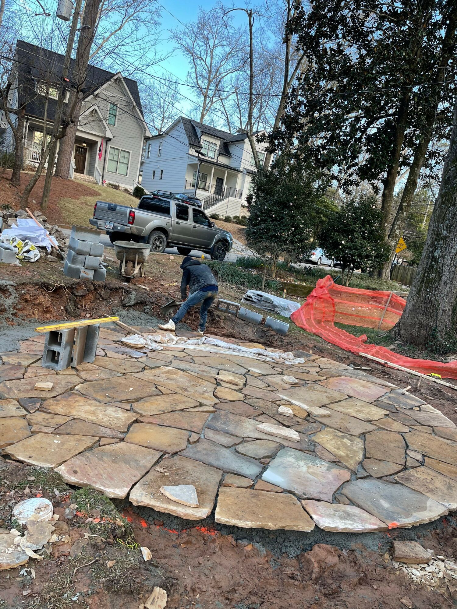 Person working on stone pathway under construction, with trees and houses in background, construction materials nearby.