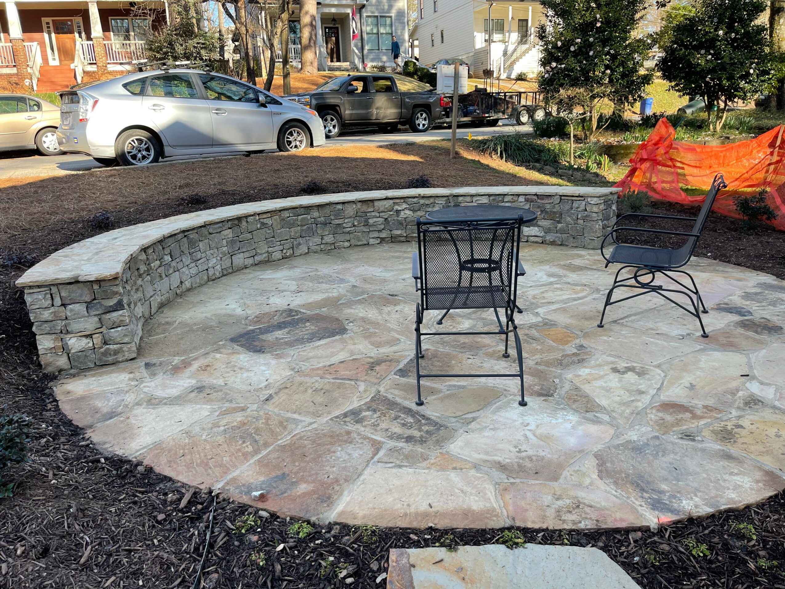 Circular stone patio with three black metal chairs, surrounded by a low stone wall, in an outdoor setting with parked cars and trees.