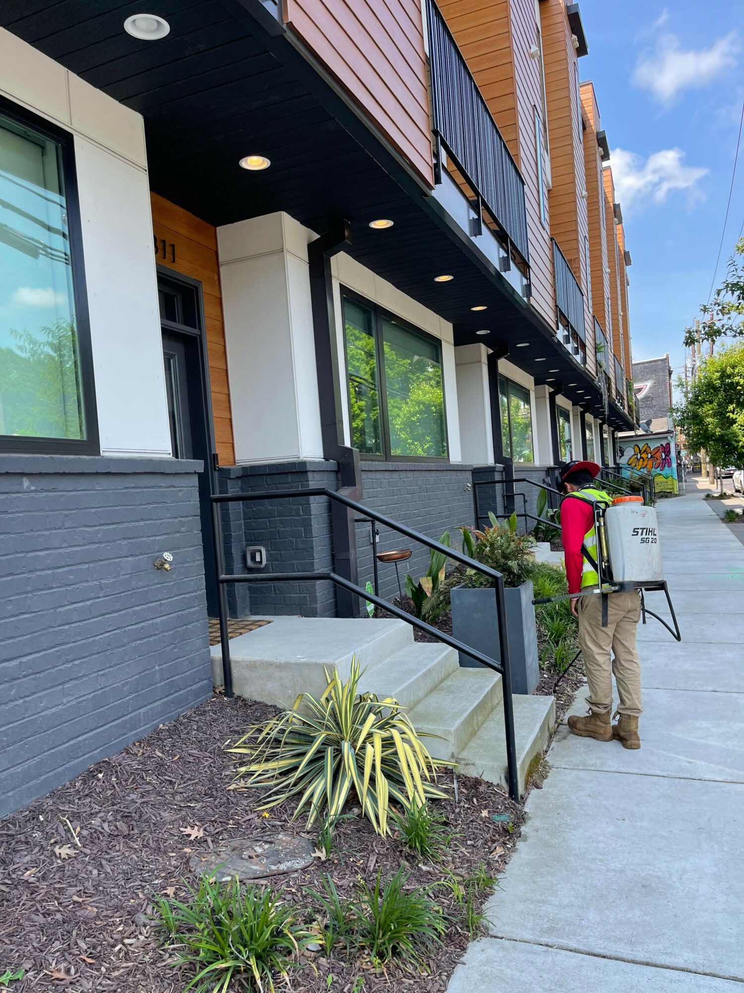 People walking on sidewalk outside modern building with large windows and outdoor seating area, plants, and a trash bin.