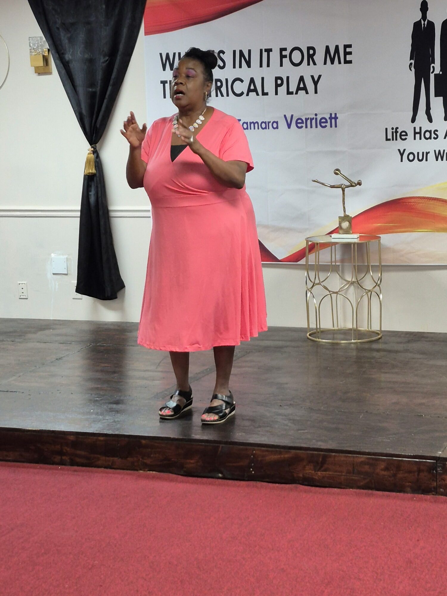 Woman in pink dress speaking on stage with a backdrop and a small table, standing on a wooden floor.
