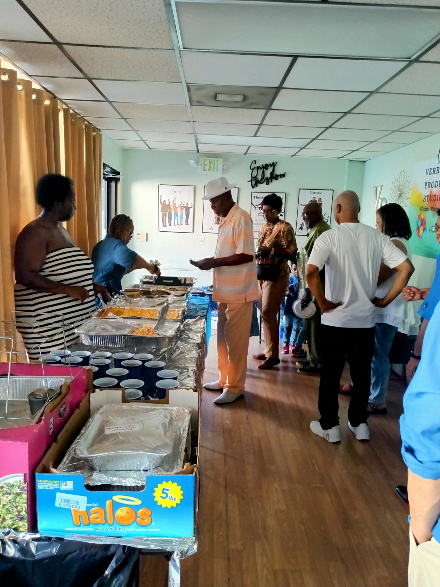 People gather around tables with food in a room with yellow curtains and a colorful wall, some standing and talking.