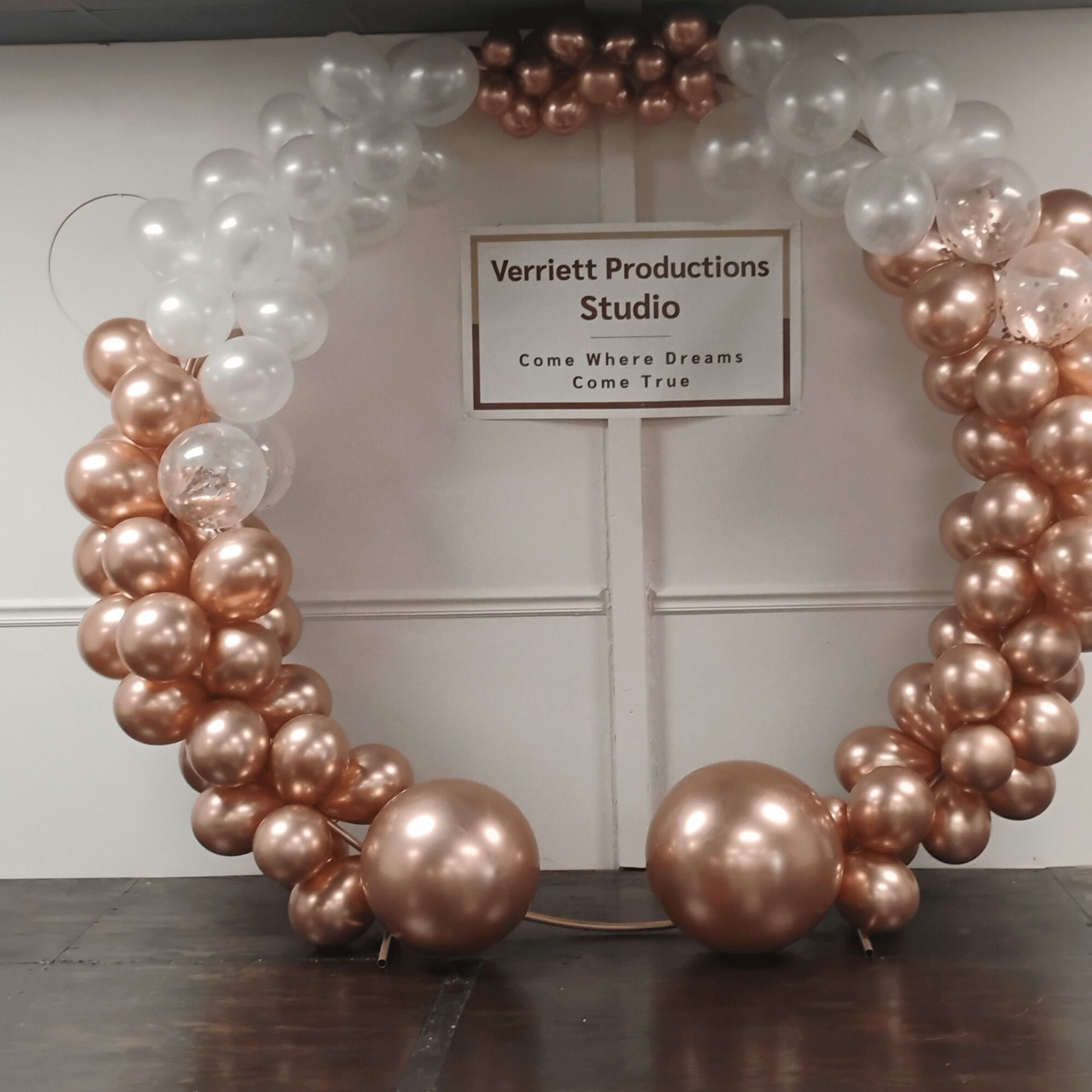 Decorative balloon arch with white and rose gold balloons, and a sign for Verriett Productions Studio, on a wall.