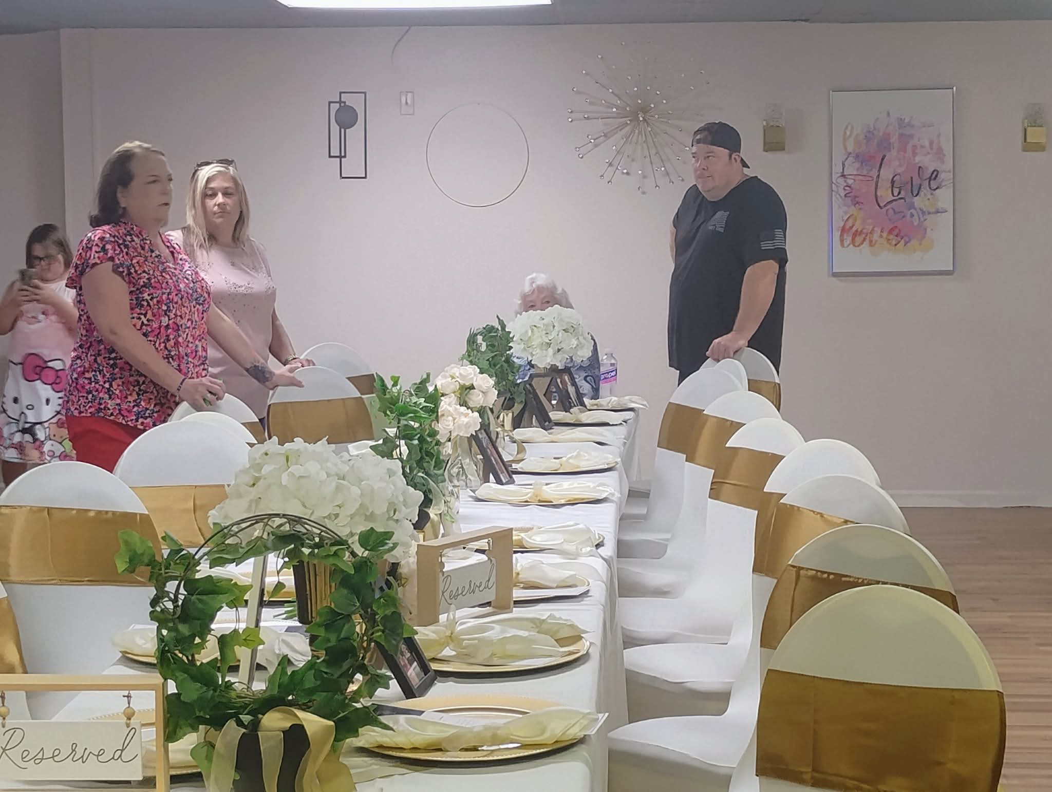 People standing around a decorated table with white chairs, flowers, and wall art in a room.