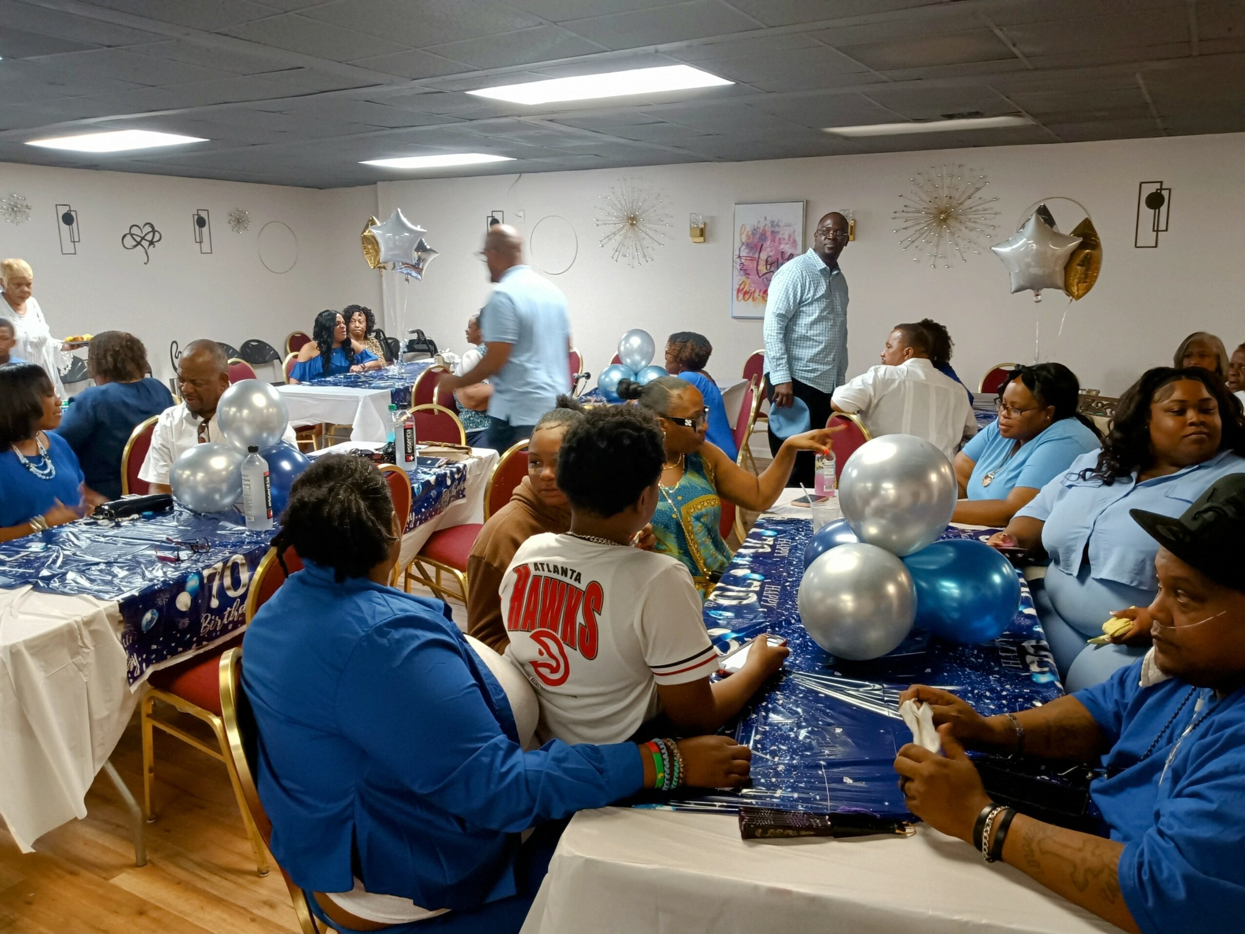 Group of people gathered in a decorated room with balloons, tables, and chairs, celebrating an event or party.