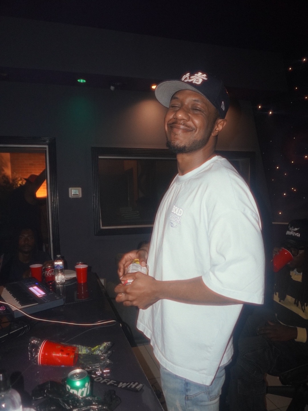 Smiling man in a white t-shirt and baseball cap holding a drink at a party, with a dark background and party supplies.