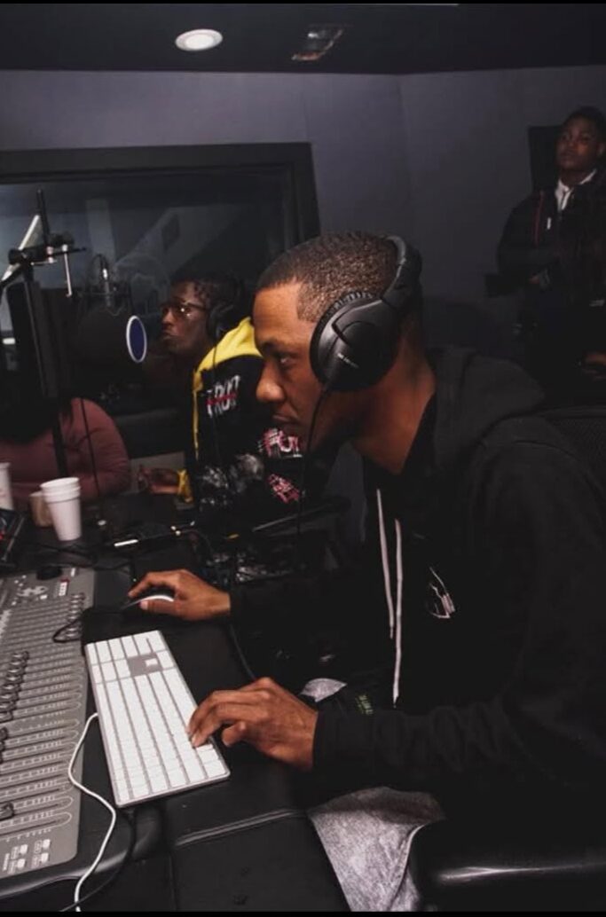 Person wearing headphones using a computer in a recording studio with others in the background.