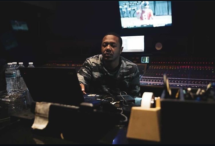 Person sitting at a mixing console in a recording studio with a monitor and water bottles nearby.