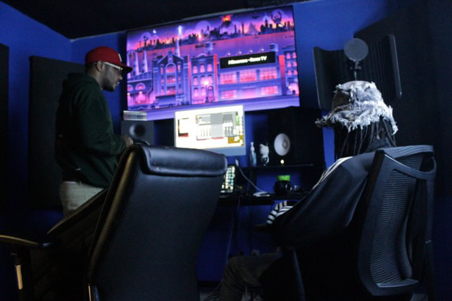 Person wearing a red cap working at a computer in a dark room with large screens and audio equipment.