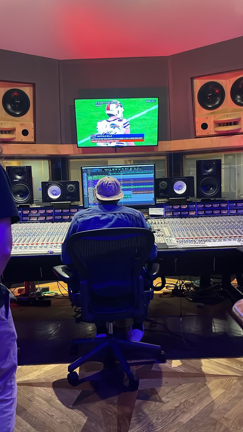 Person sitting in front of a mixing console and speakers in a recording studio, watching a TV screen showing a sports game.