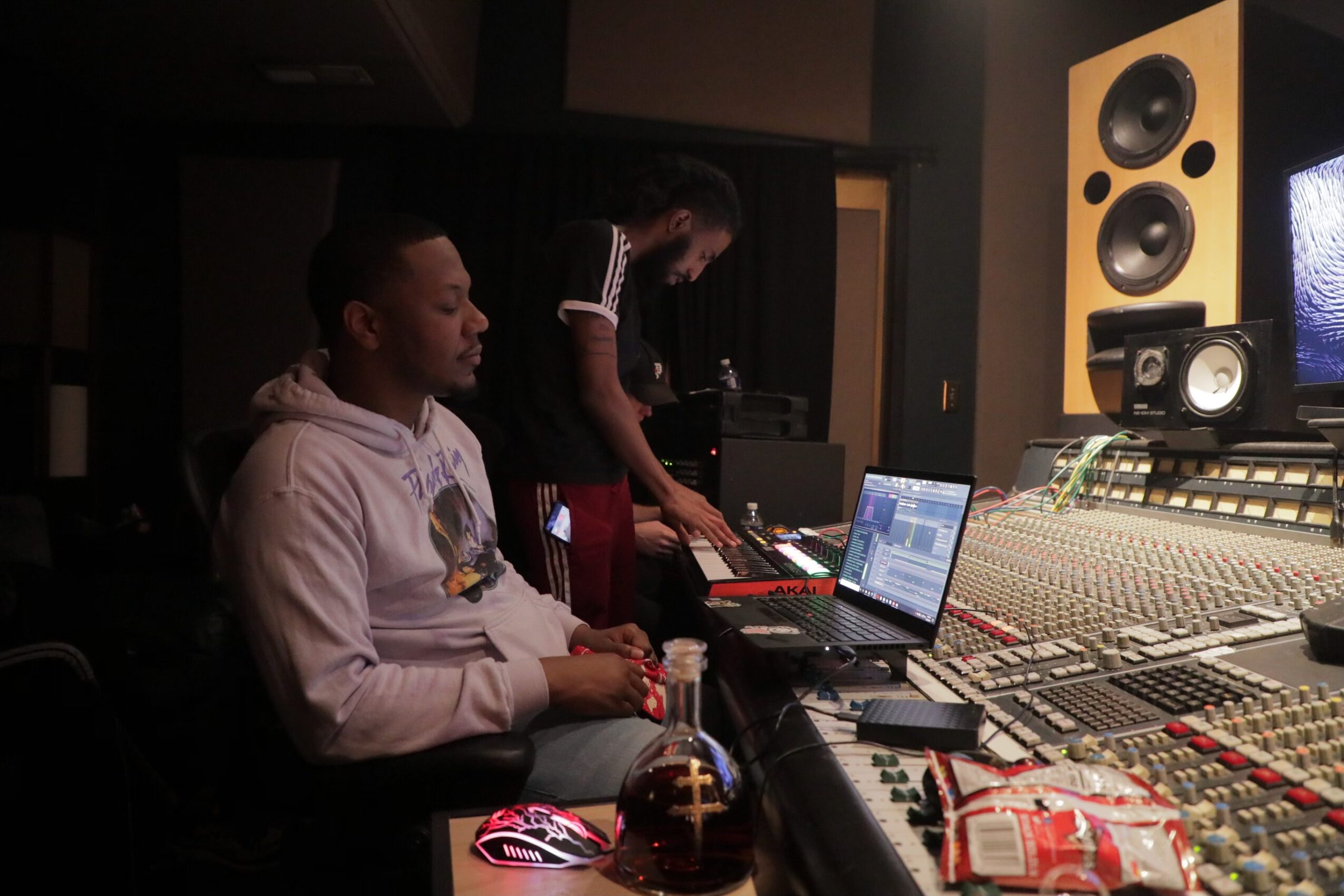 Two people in a recording studio with audio equipment and speakers, one sitting and one standing, working on a laptop.