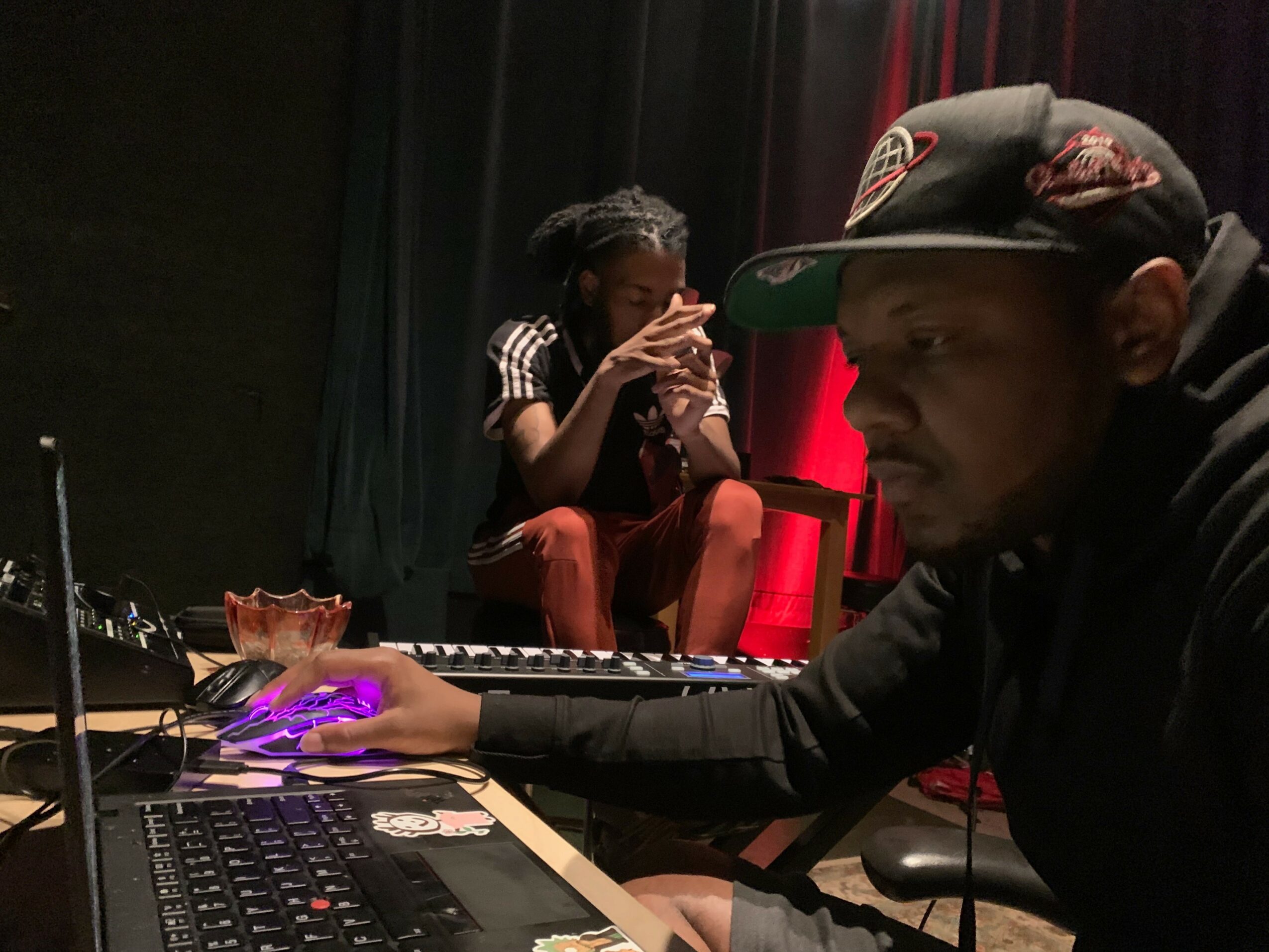 Two people in a dimly lit room with a keyboard and electronic equipment, one person wearing a cap and the other sitting on a chair.