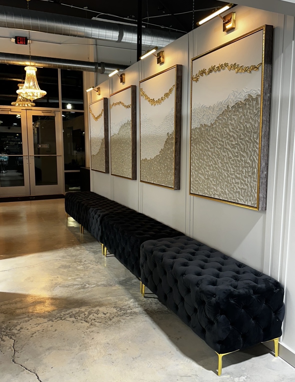 Interior with four framed landscape artworks on wall, black tufted bench below, chandelier visible through glass door, and polished floor.
