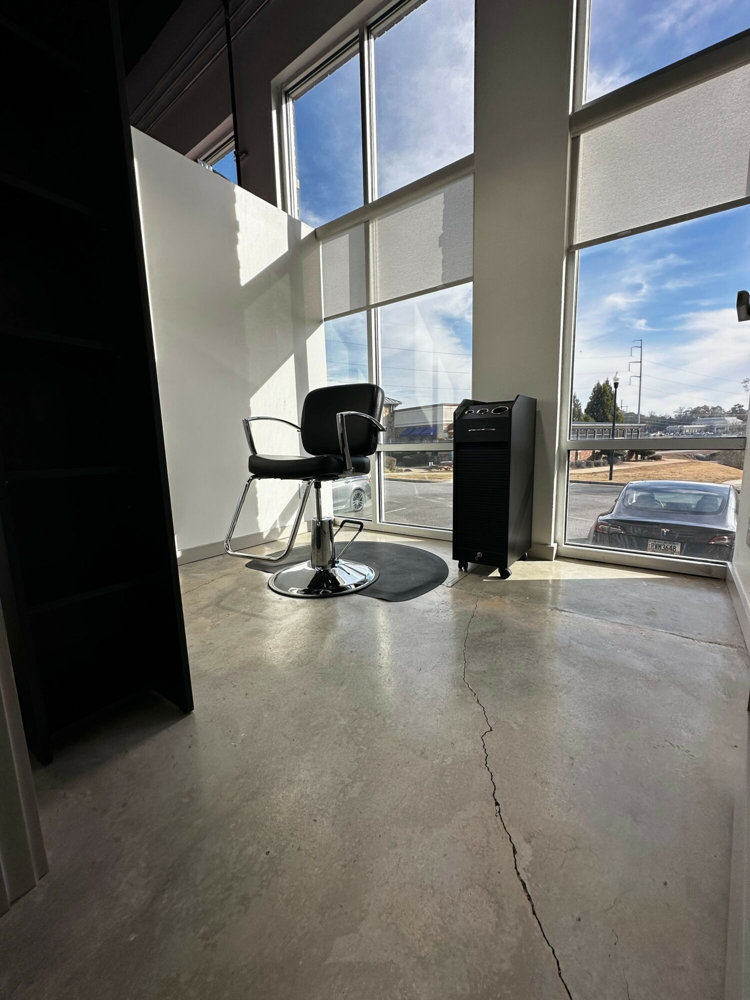Interior view of a room with large windows showing a parking lot and blue sky, a black chair, and a small table.