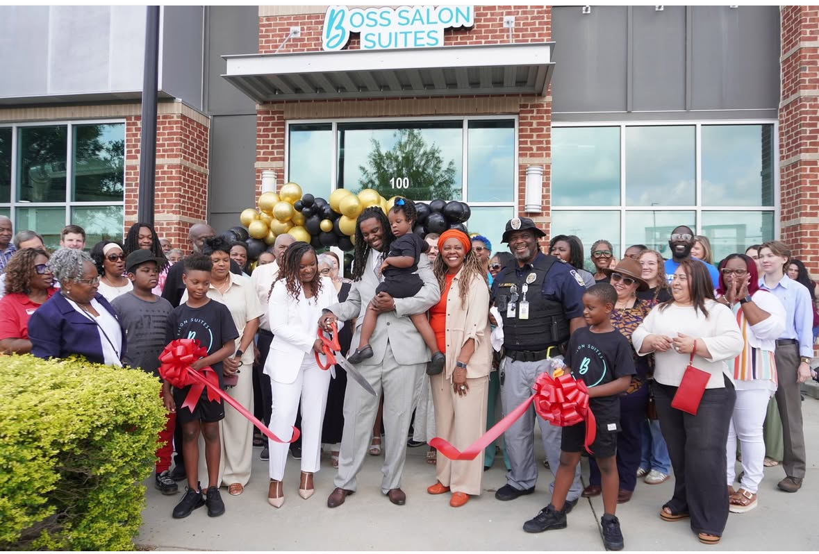 Group of people gathered outside a building for a ribbon-cutting event, with balloons and a red ribbon, celebrating opening.