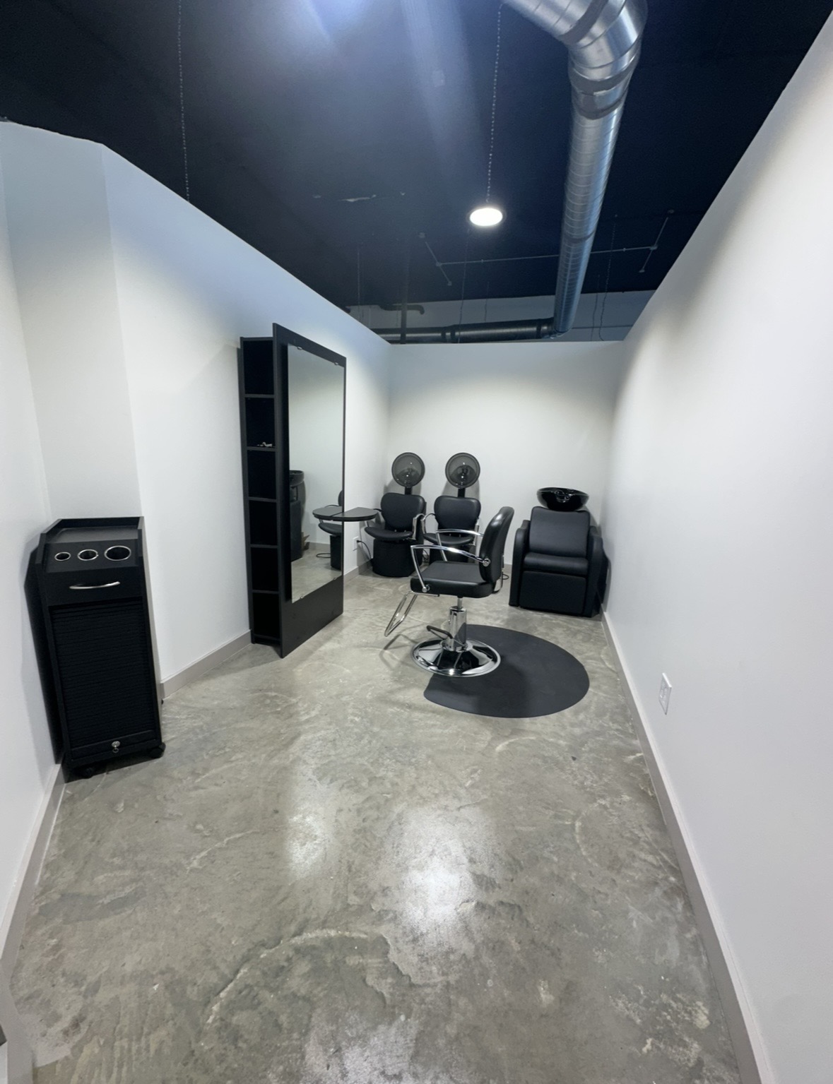 Salon or barber shop with chairs, mirror, and equipment in a narrow room, concrete floor, white and black walls.