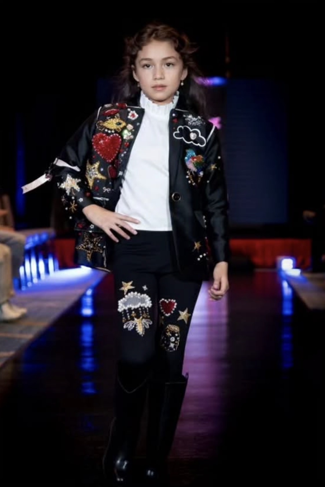 Child walking on runway wearing a decorated jacket and pants with patches and designs, dark background, illuminated runway